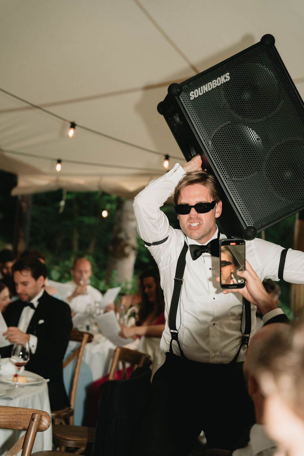 A person in formal attire and sunglasses dances energetically under a white tent at a wedding reception.