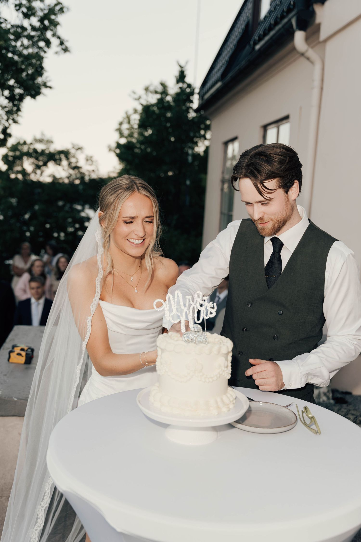 Bride in strapless gown and veil with groom in vest cutting white wedding cake at outdoor reception.