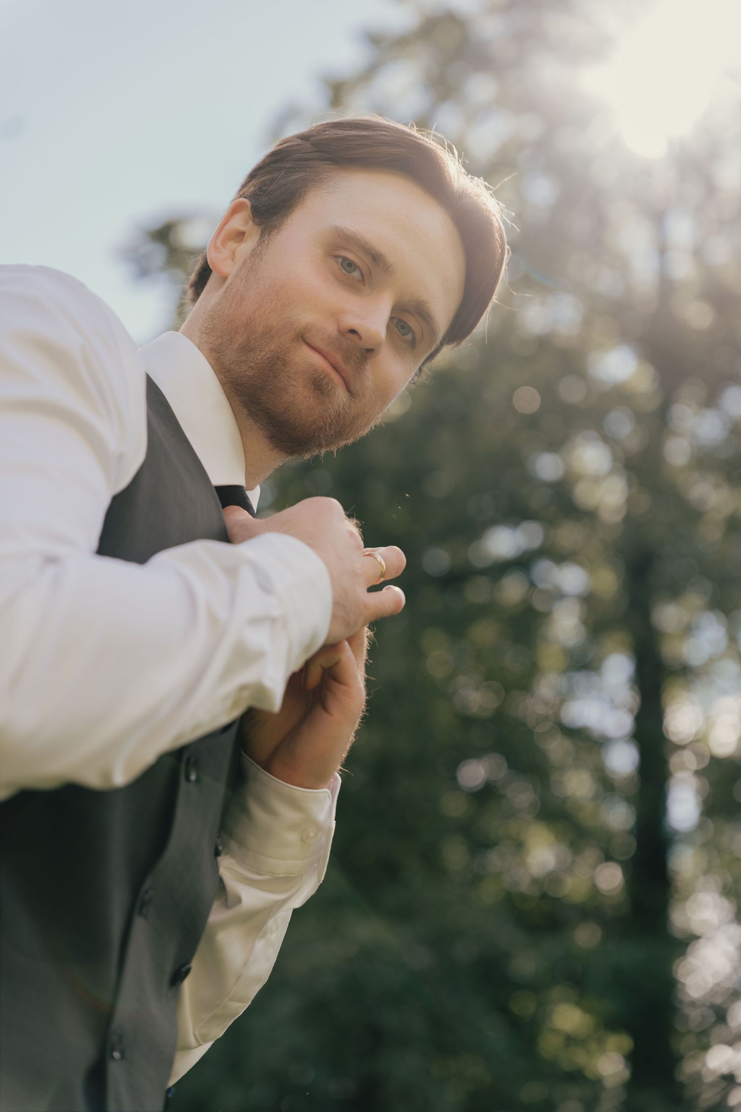 Dapper groom in vest and tie adjusting his outfit outdoors in golden sunlight.