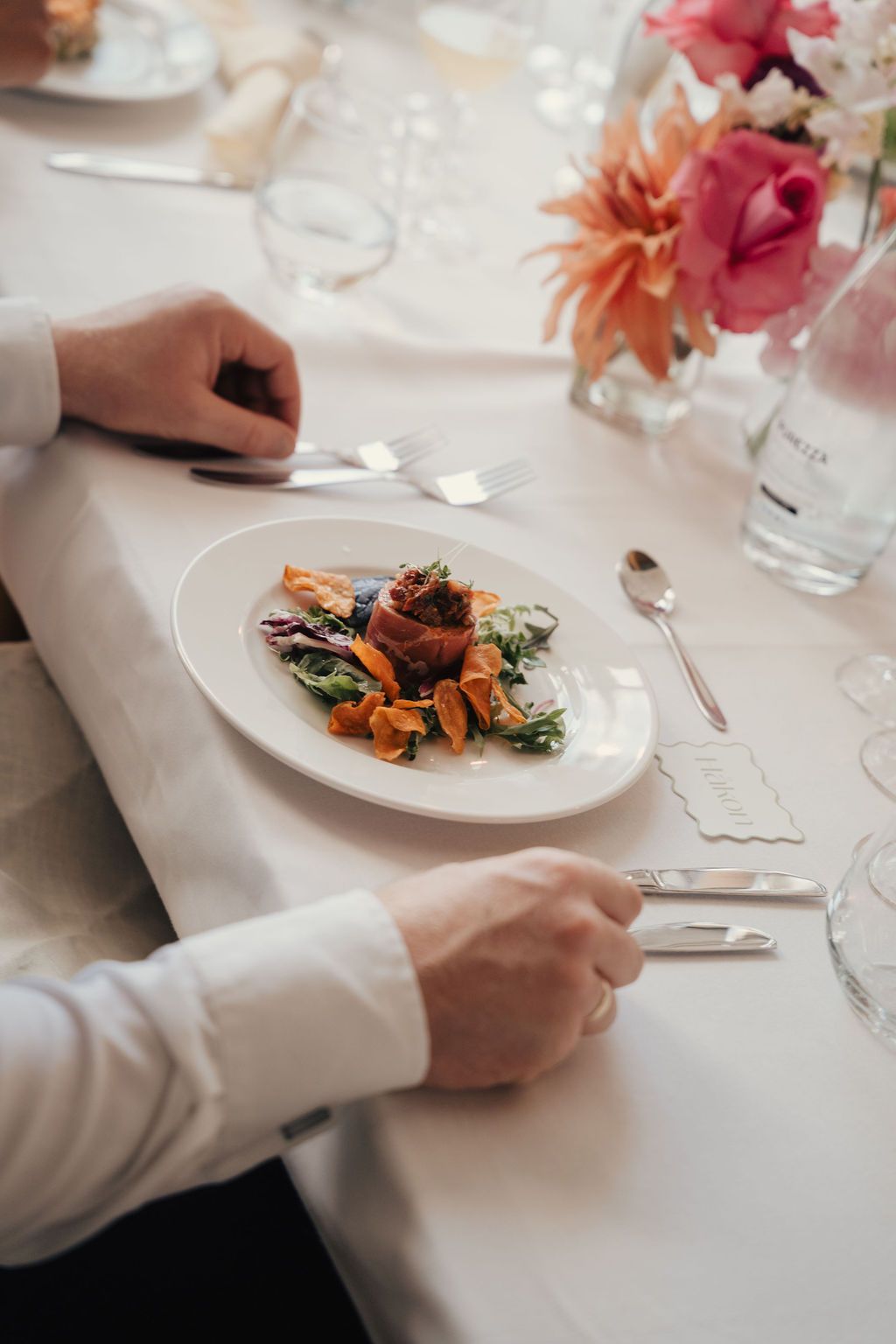 Elegant table setting at upscale restaurant with pink flowers and hands reaching for plated gourmet dishes.