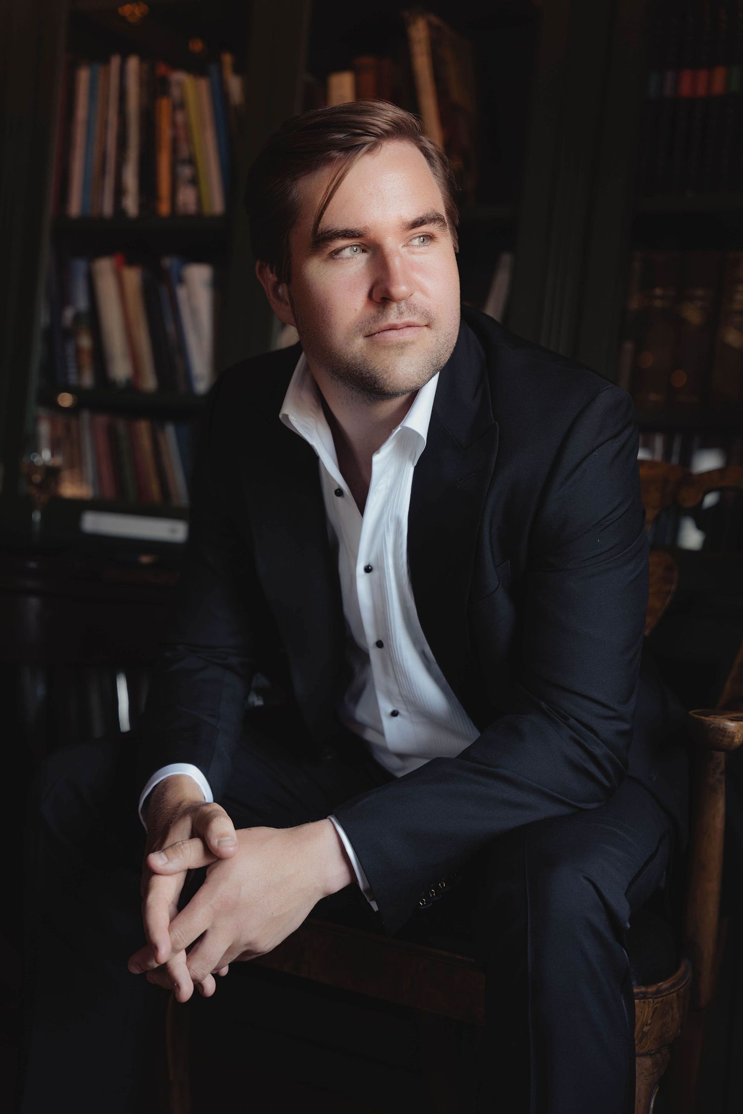 A man in a dark suit sits thoughtfully in a library chair with bookshelves in the background.