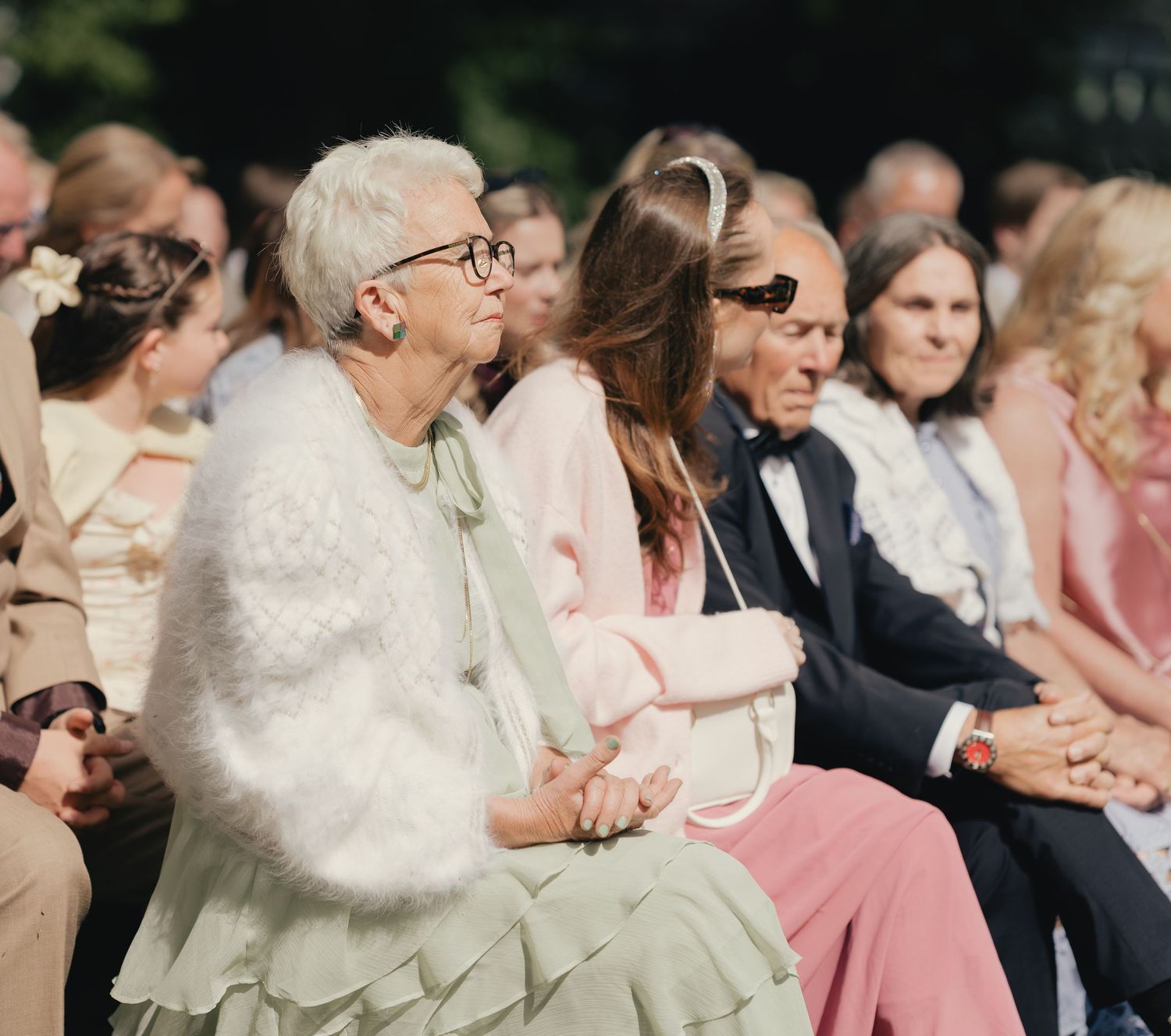 Elegantly dressed wedding guests seated outdoors, including an older woman in a white fuzzy cardigan and green dress.
