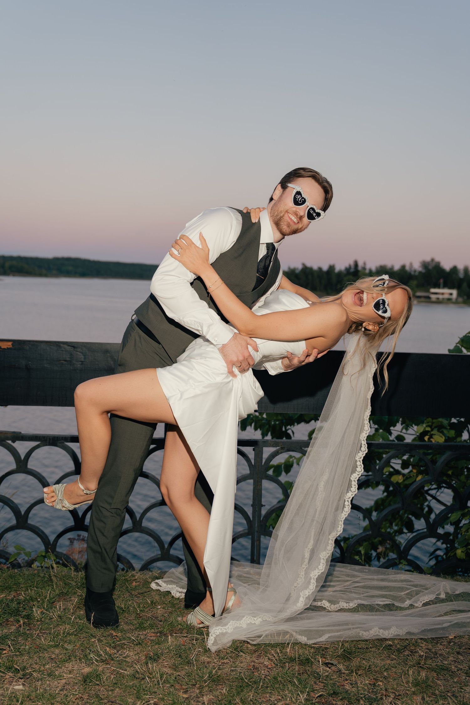 Groom dips bride in white dress and veil at sunset near waterfront railing.