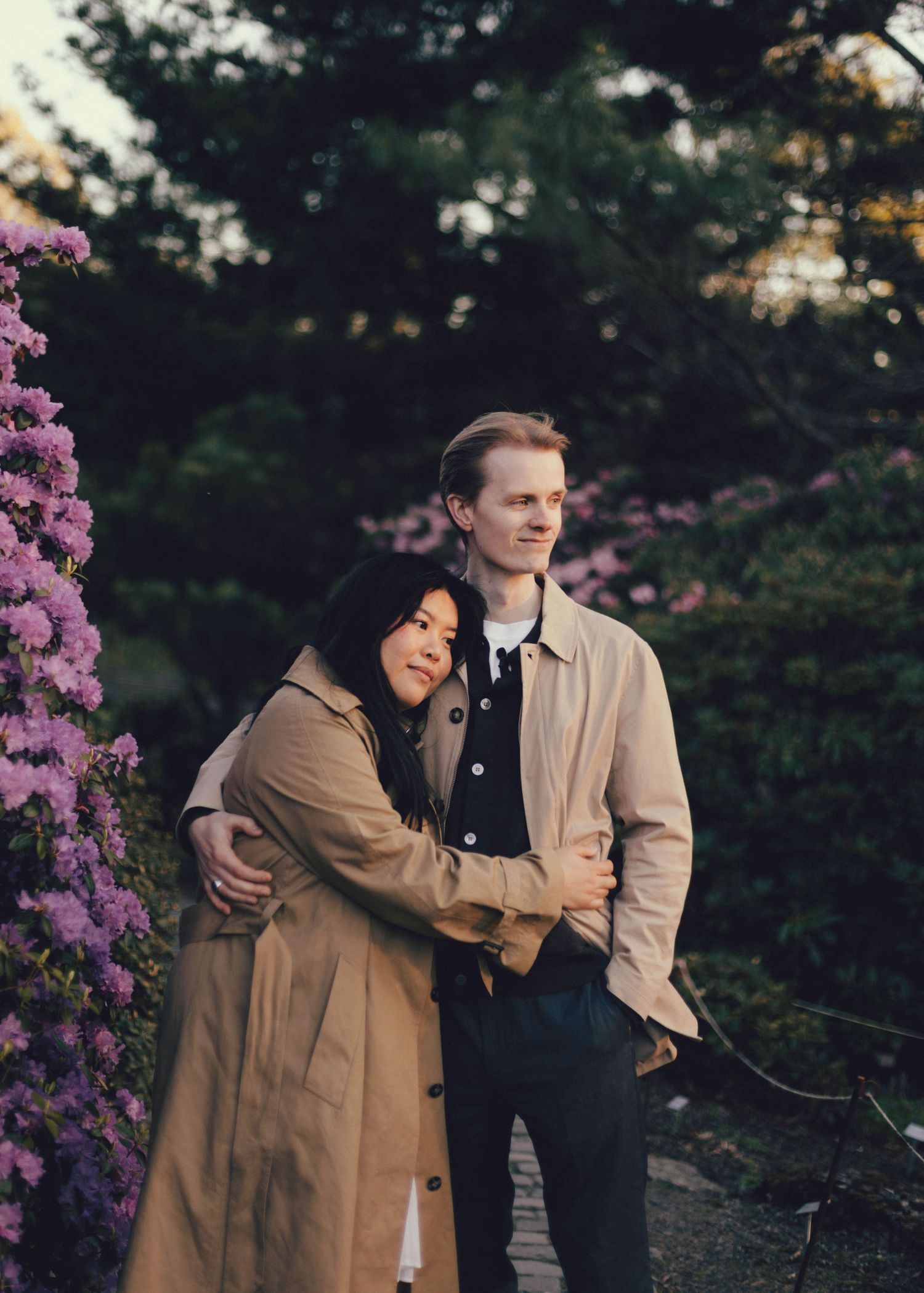 A couple in matching beige coats embrace while standing in a garden with blooming purple flowers.