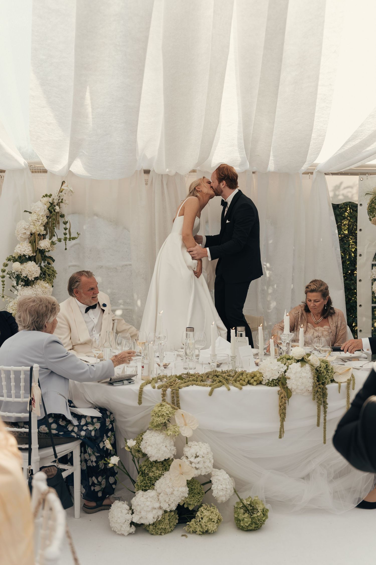 Bride and groom share a kiss beside a white-draped reception table adorned with white flowers while seated guests look on.