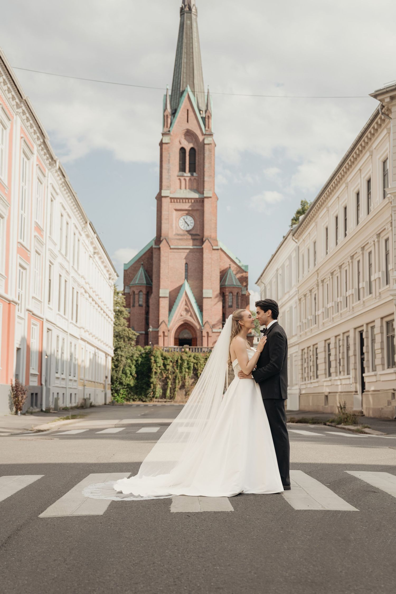 Bride and groom embrace on a crosswalk in front of a red brick church with a tall spire, framed by pastel historic buildings.