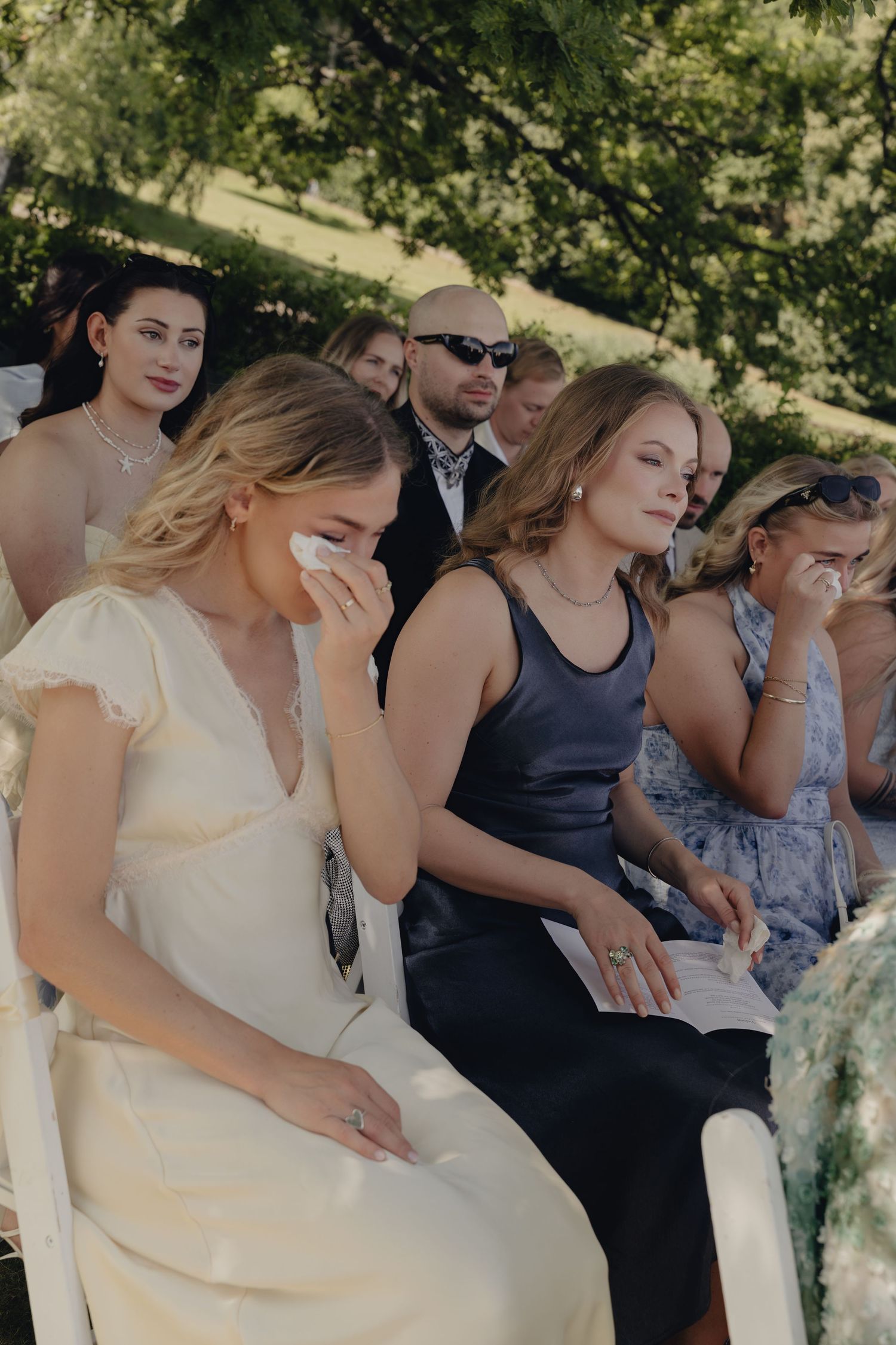 Emotional wedding guests wipe away tears during an outdoor ceremony seated on white chairs beneath leafy trees.
