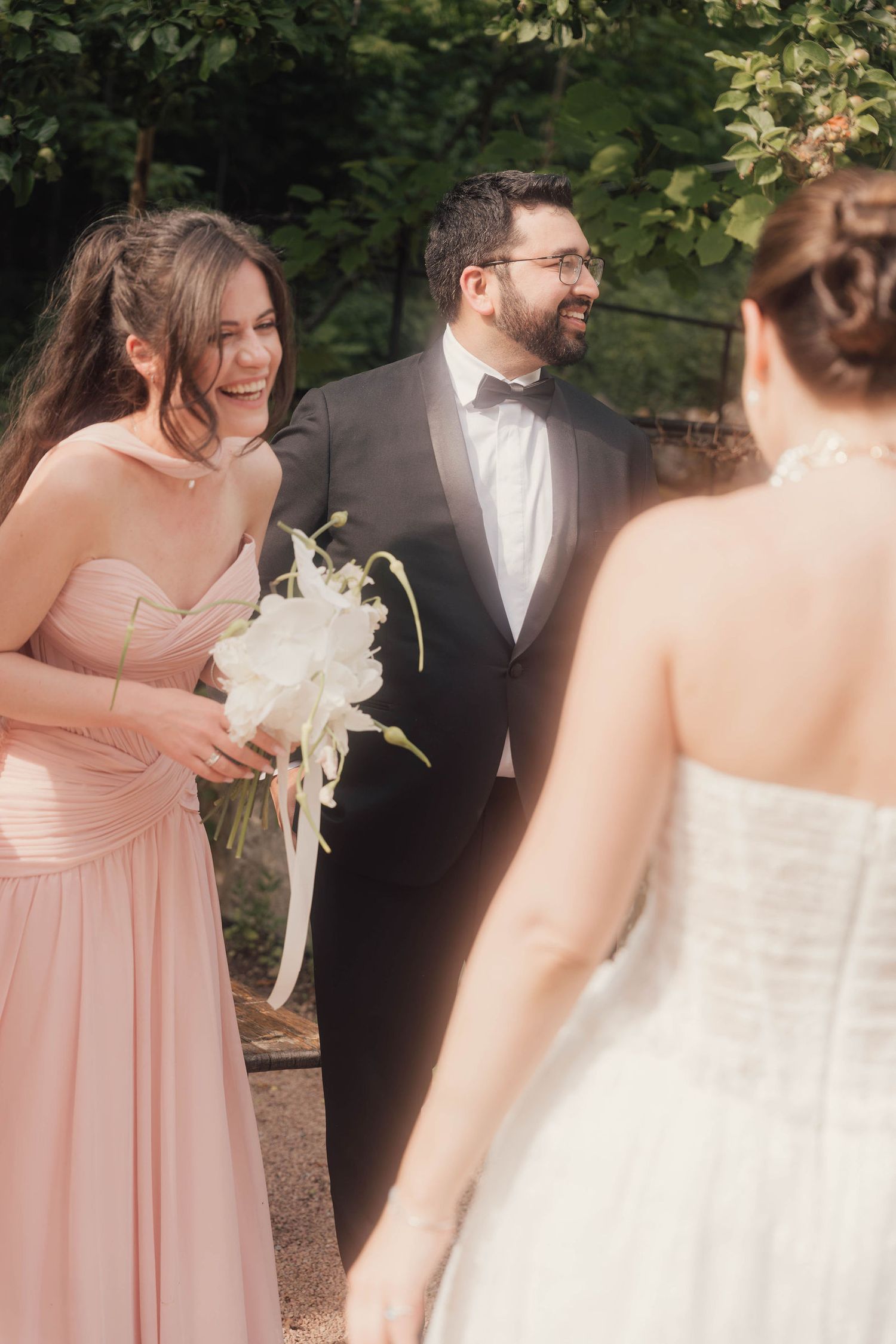 Bride in white gown greets smiling bridesmaid in pink dress and groom in black tuxedo at outdoor wedding ceremony.
