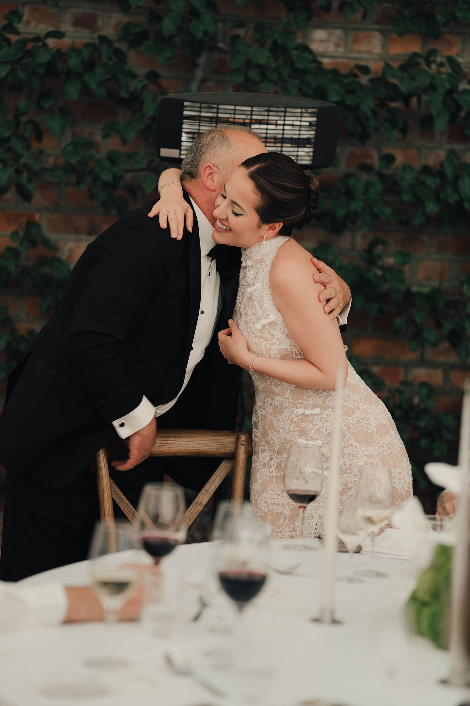 Bride and older gentleman share a tender embrace at a wedding reception table with red wine glasses and ivy backdrop.