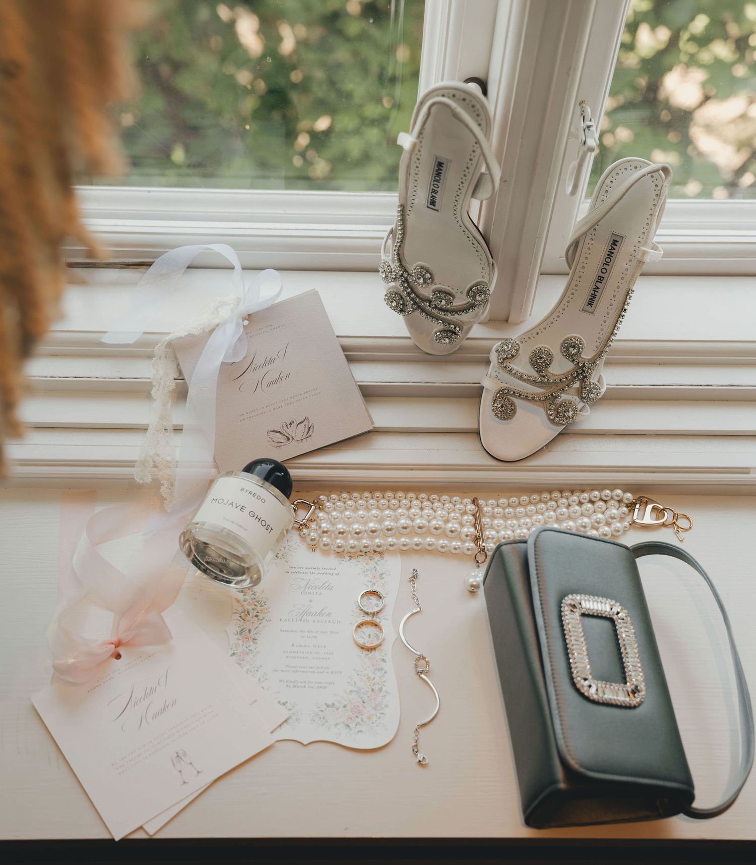 Elegant bridal flat lay featuring jeweled heels, pearl bracelet, gray clutch, gold rings, perfume, and wedding invitations on white surface.