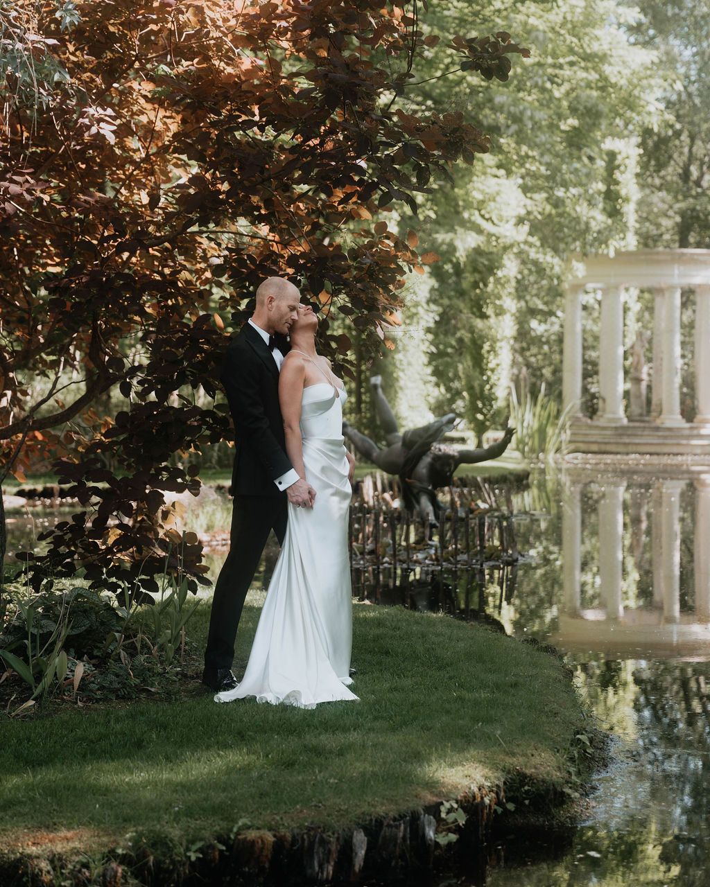 A couple shares a romantic moment in a lush garden setting with ornate stone columns and greenery.