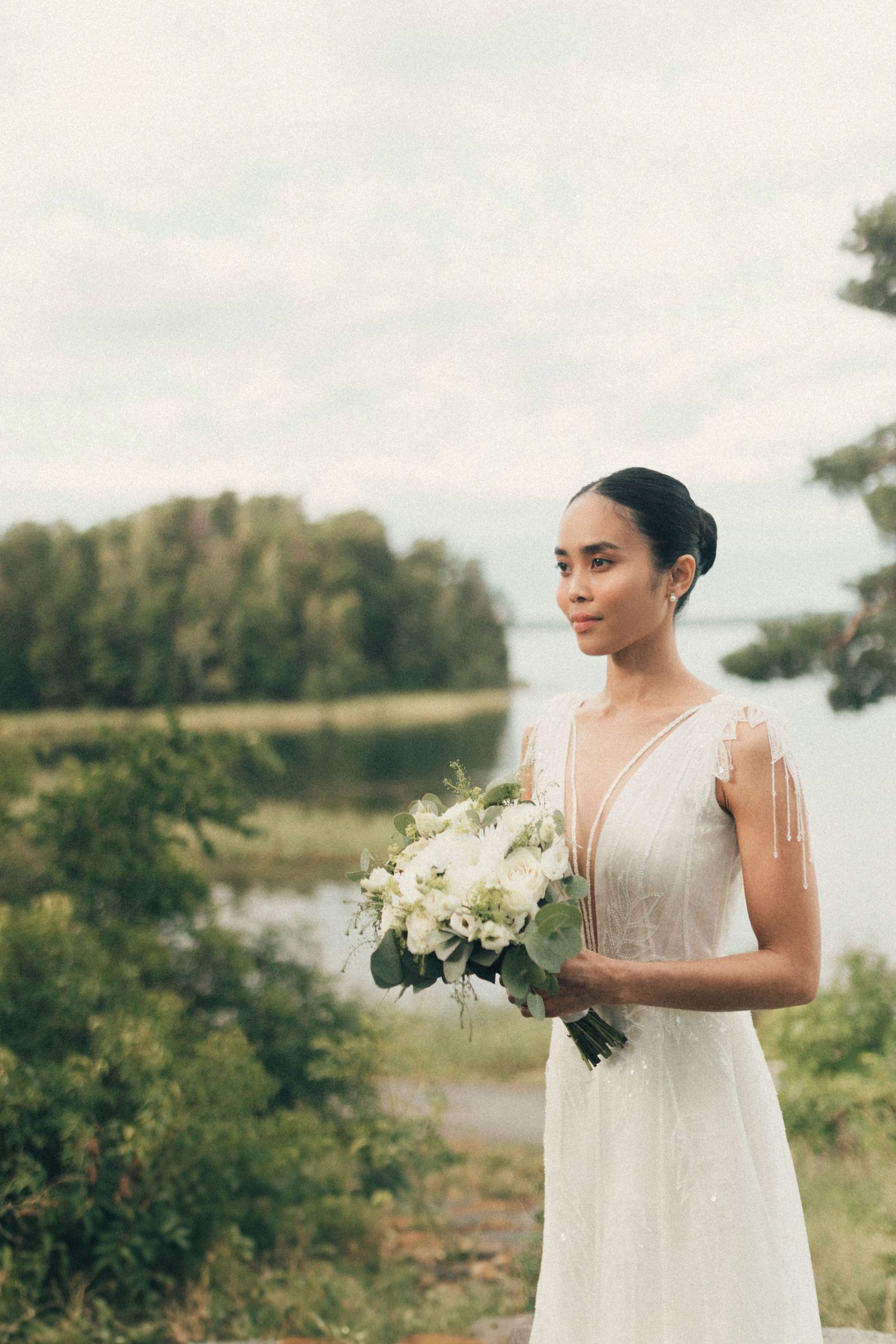 A bride in a white dress holds a delicate bouquet of white flowers against a serene outdoor landscape.