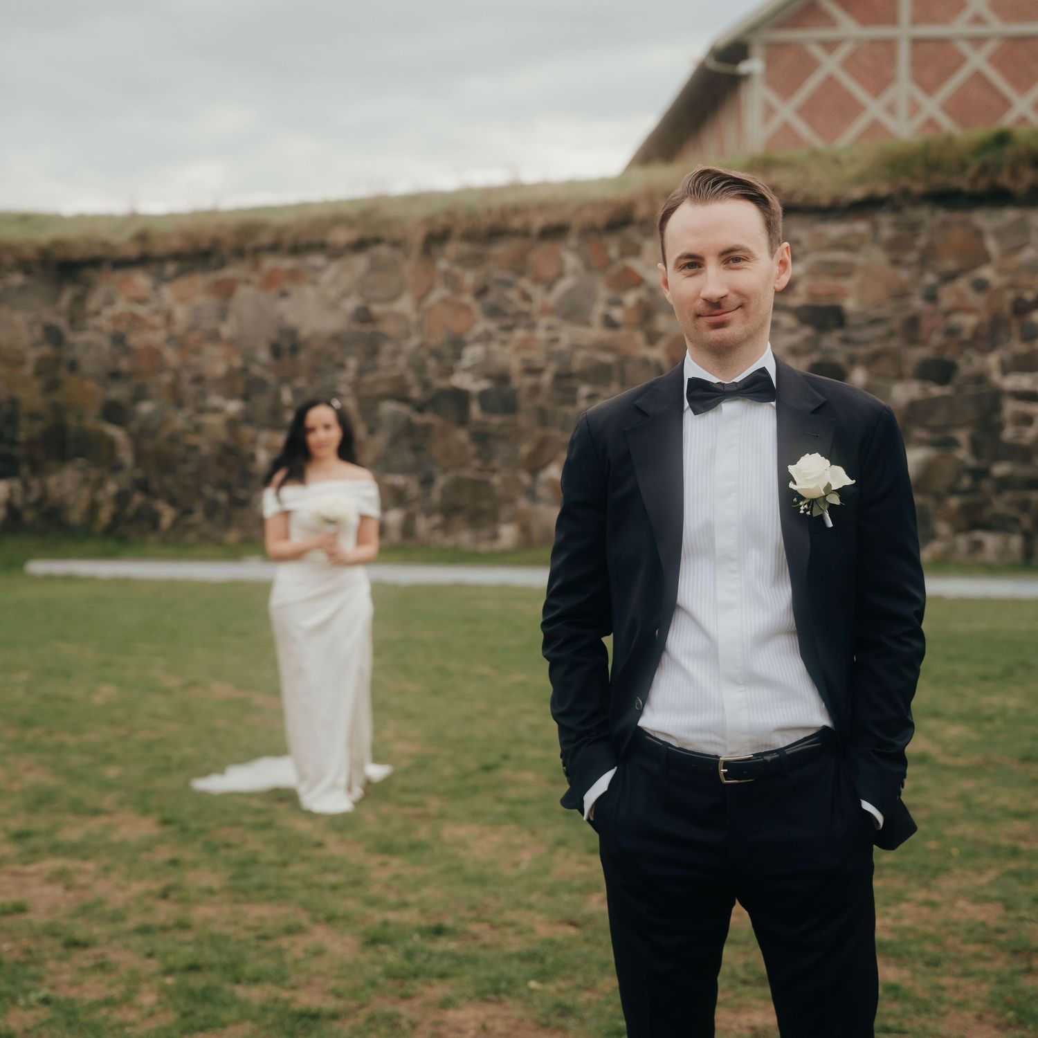 A couple in wedding attire poses against a rustic stone wall backdrop in an outdoor setting.