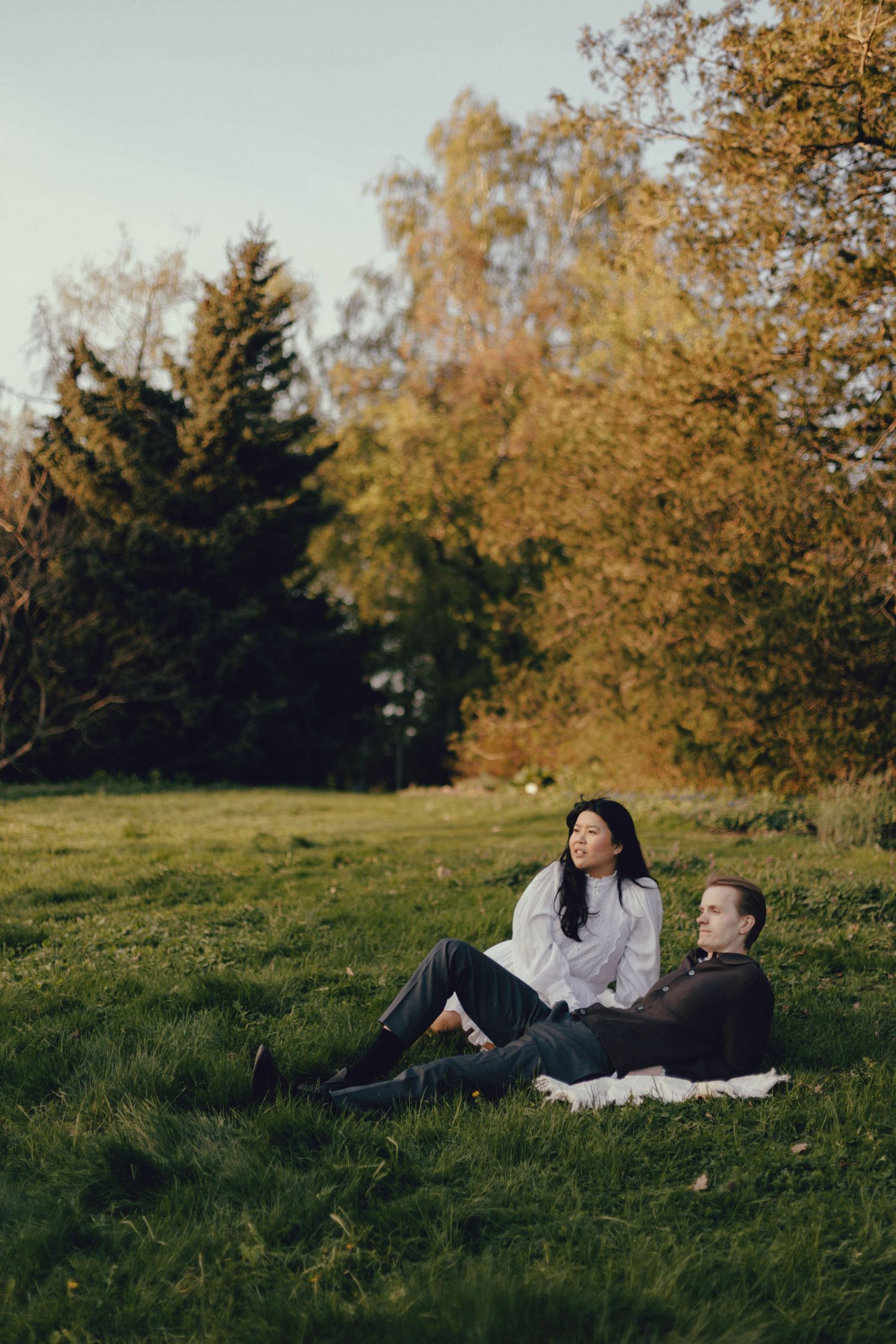 Couple relaxing together on grassy hill during autumn afternoon with trees in background.