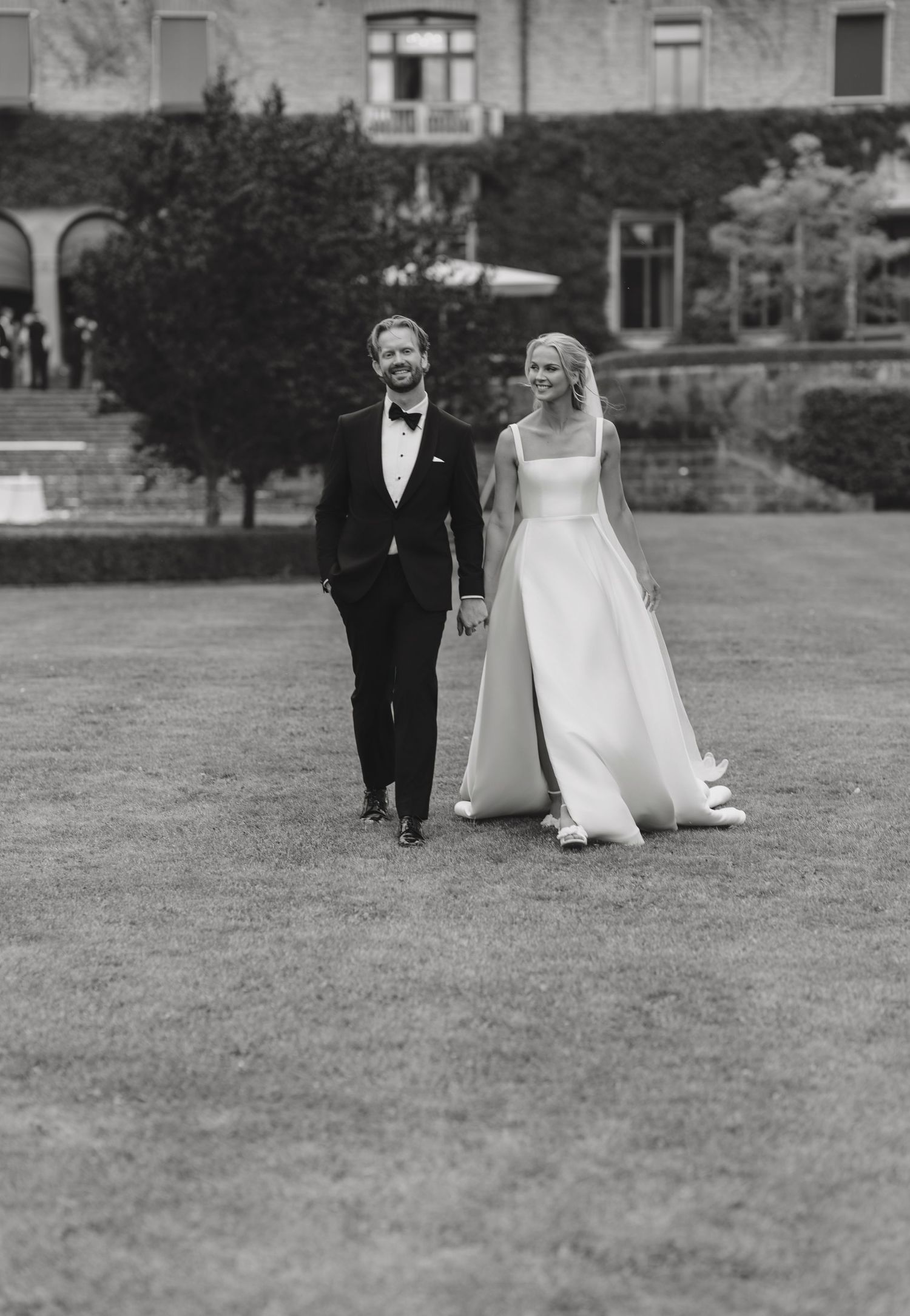 Smiling bride in a sleek strapless gown holds hands with a groom in a tuxedo and bow tie while walking across a lawn at a historic estate.