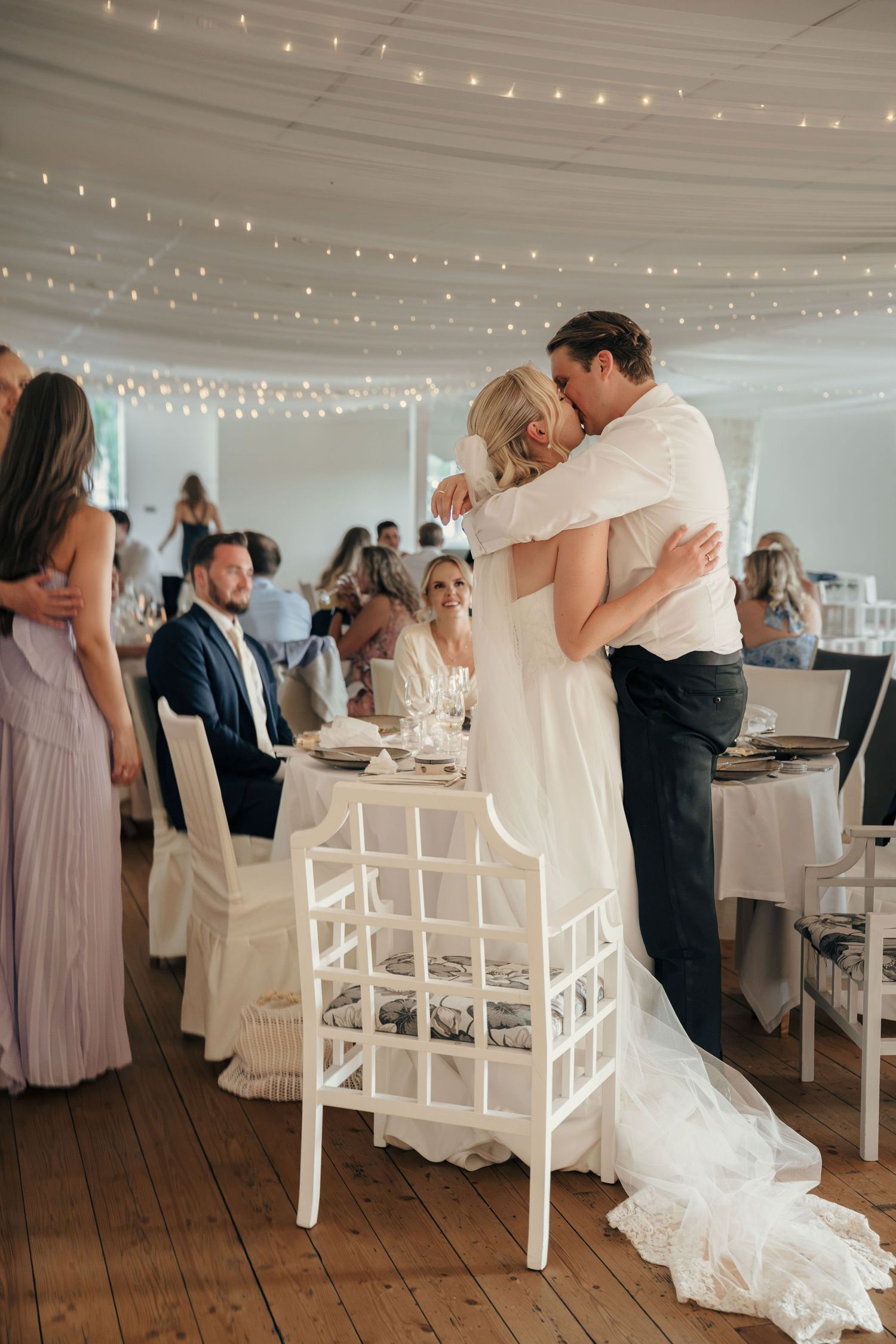 A bride and groom share an embrace beside a white chair in a tent reception hall with string lights and seated wedding guests.
