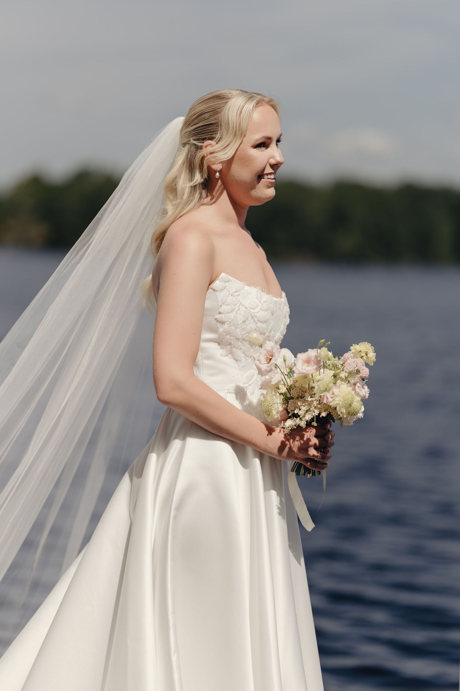 Smiling bride in a strapless white satin gown and long veil holds a pastel bouquet beside a lake on a sunny day.