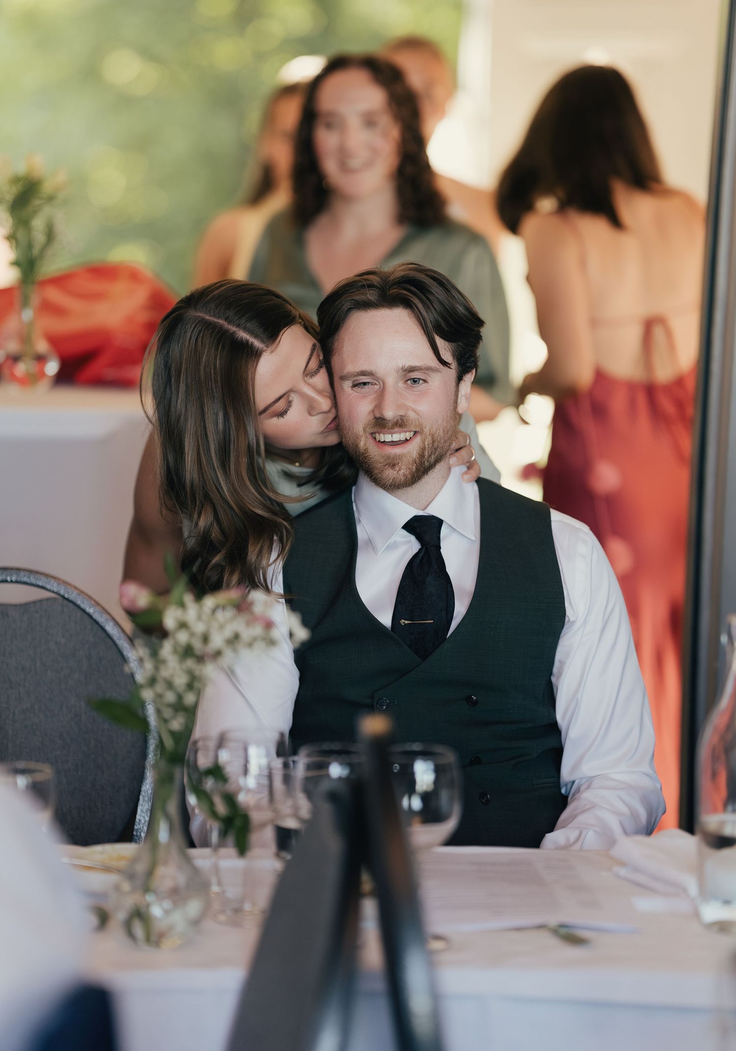 Couple sharing a tender moment at wedding reception table with guests celebrating in background.