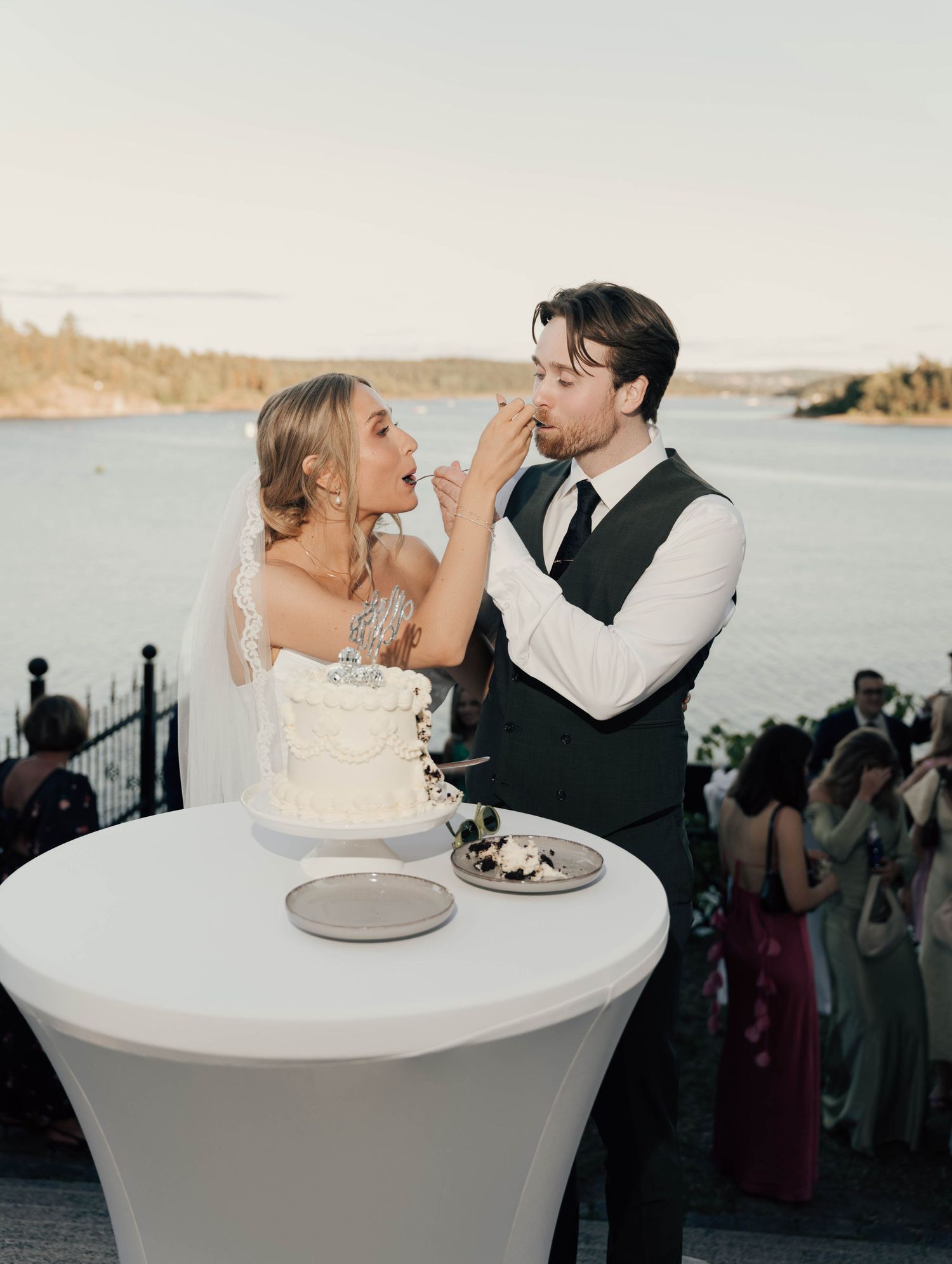Bride and groom cutting wedding cake outdoors by a scenic lake at sunset.