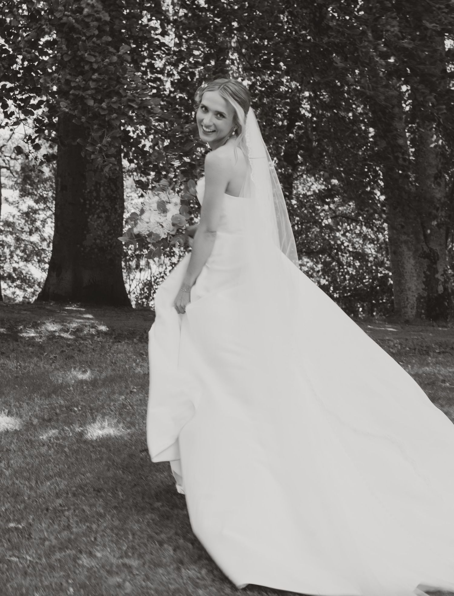 Black and white portrait of smiling bride in white gown and veil standing among trees outdoors.