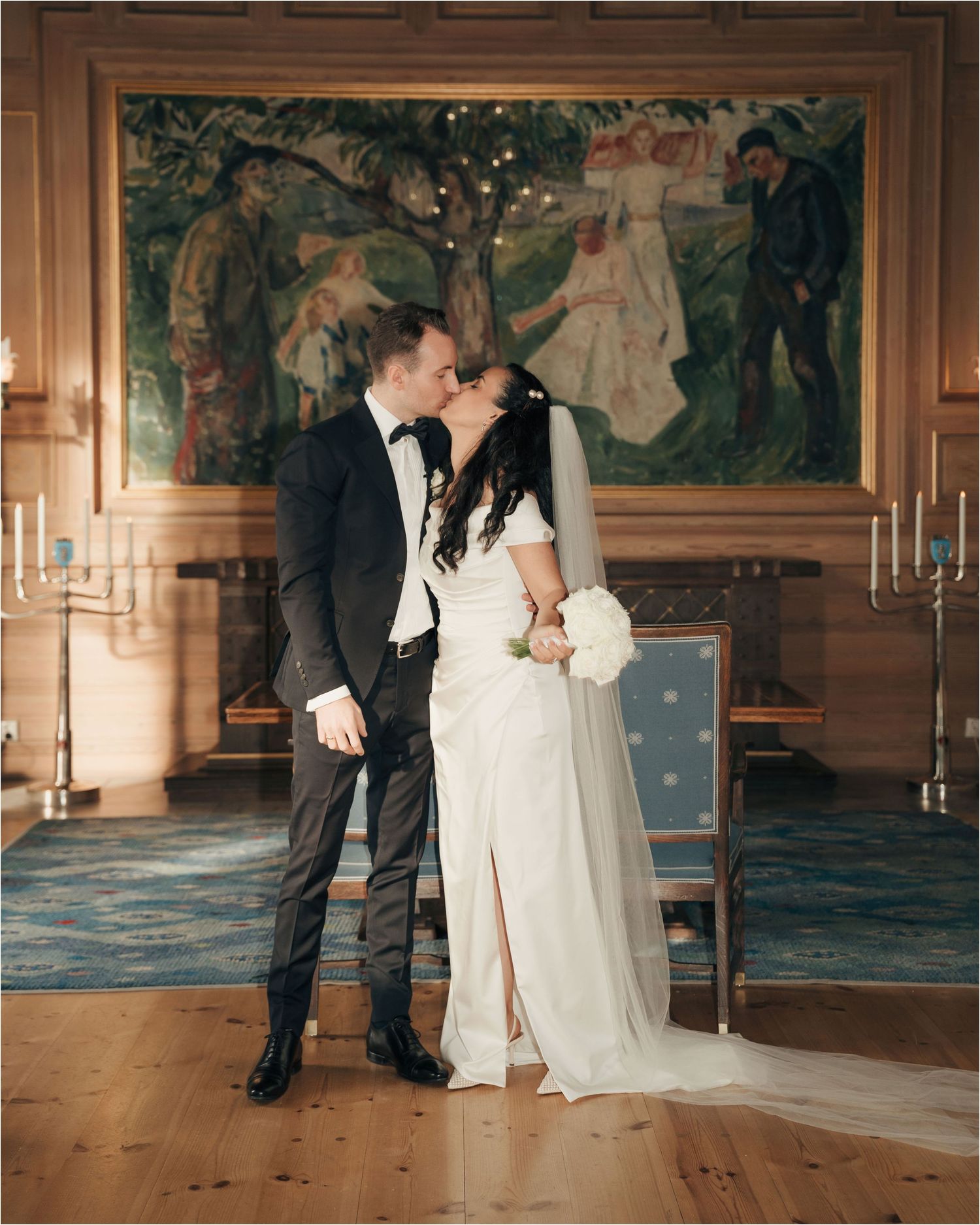 A romantic wedding moment in an elegant room with a large decorative wall mural and candlesticks.