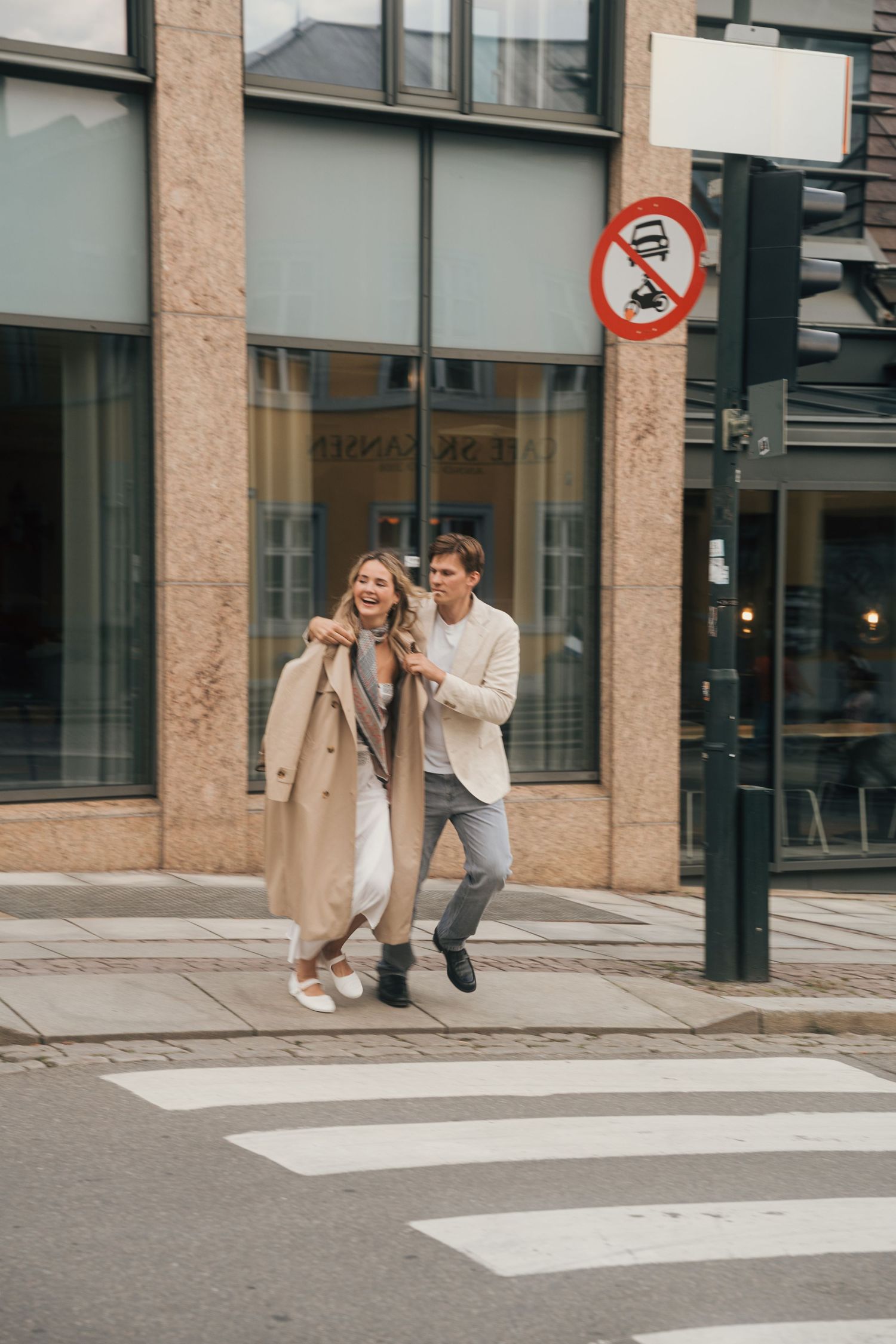 A couple playfully runs and chases each other across a pedestrian crossing in an urban setting.