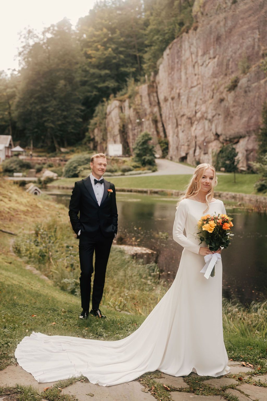 A couple in formal wedding attire poses near a scenic lake with dramatic rock cliffs in the background.