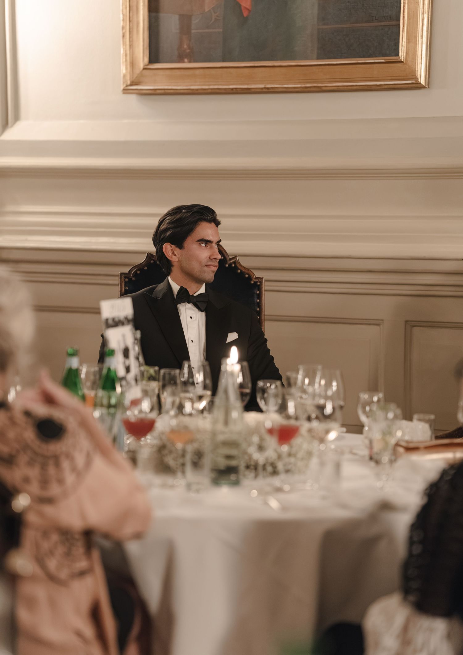 A man in a black tuxedo and bow tie sits at a candlelit banquet table with wine glasses beneath a gilded framed painting.