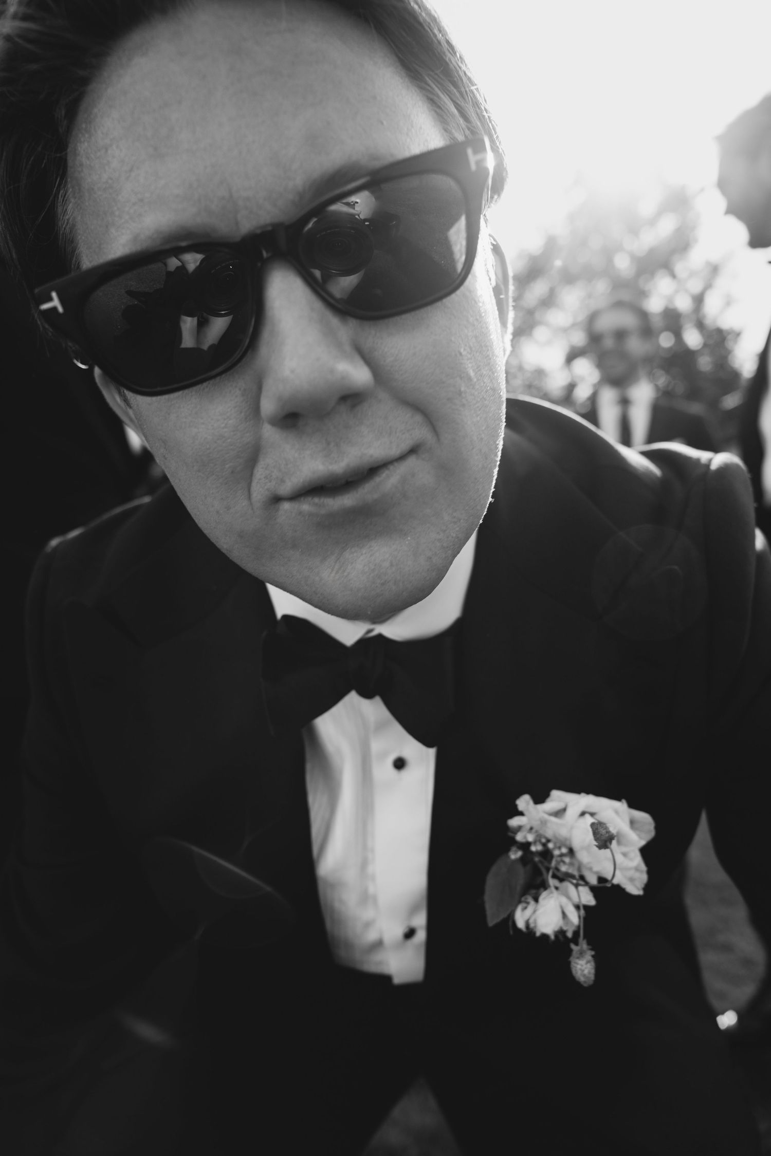 Black and white close-up of a groom in sunglasses, tuxedo and bow tie with a floral boutonniere, guests blurred behind him.