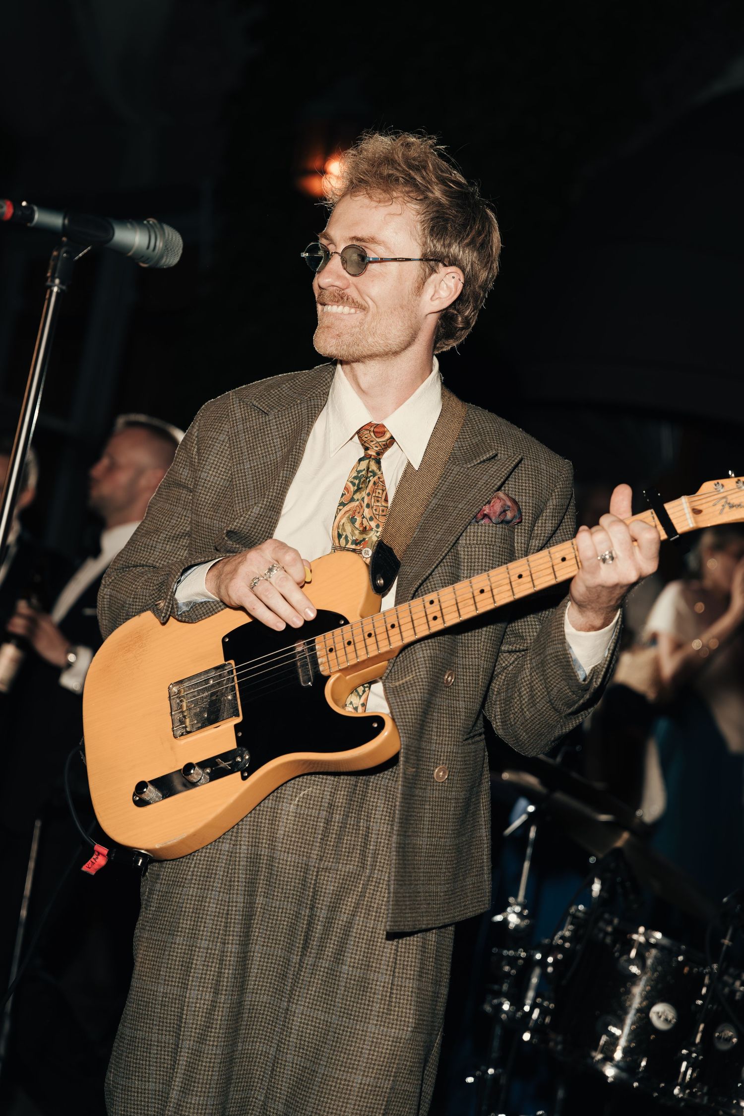 Smiling guitarist in round sunglasses, a plaid suit and patterned tie plays a butterscotch electric guitar on stage at a wedding.