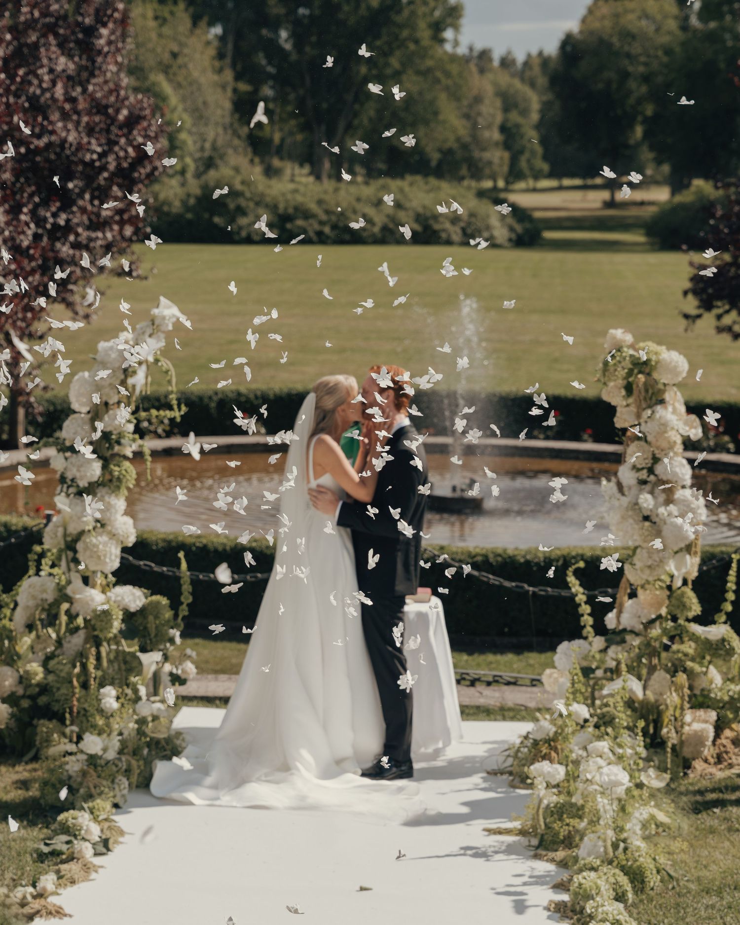 Bride and groom kiss on a white aisle runner as petals fall, surrounded by white floral arrangements in a garden.