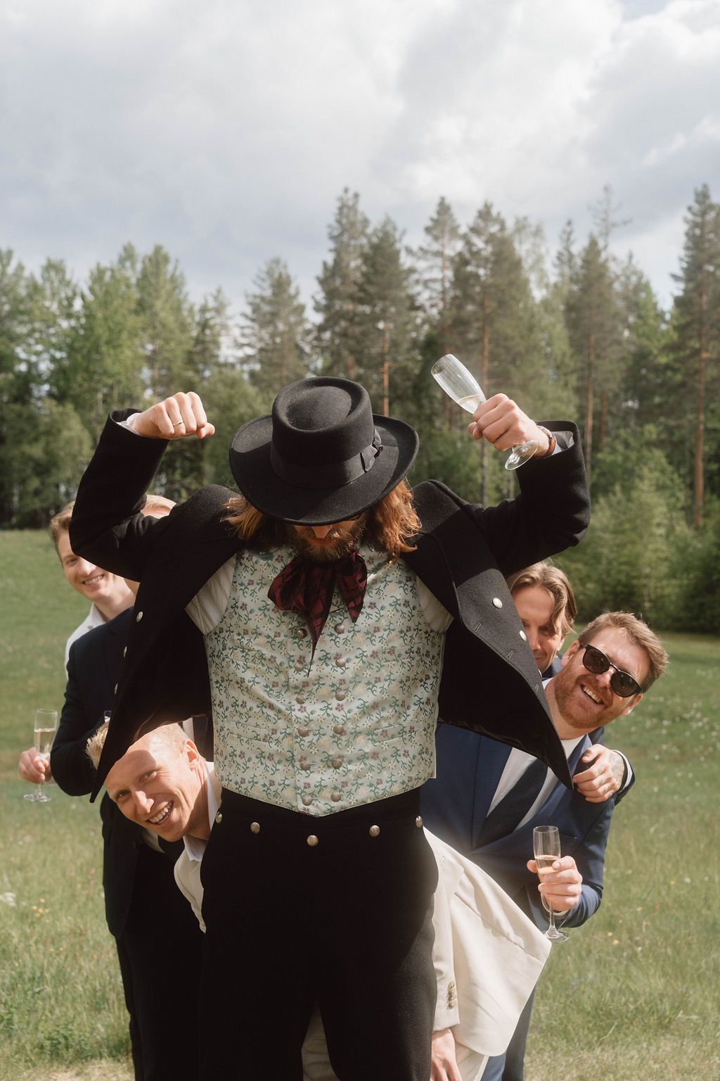 Wedding party members in cowboy attire pose playfully in a grassy field with pine trees in the background.