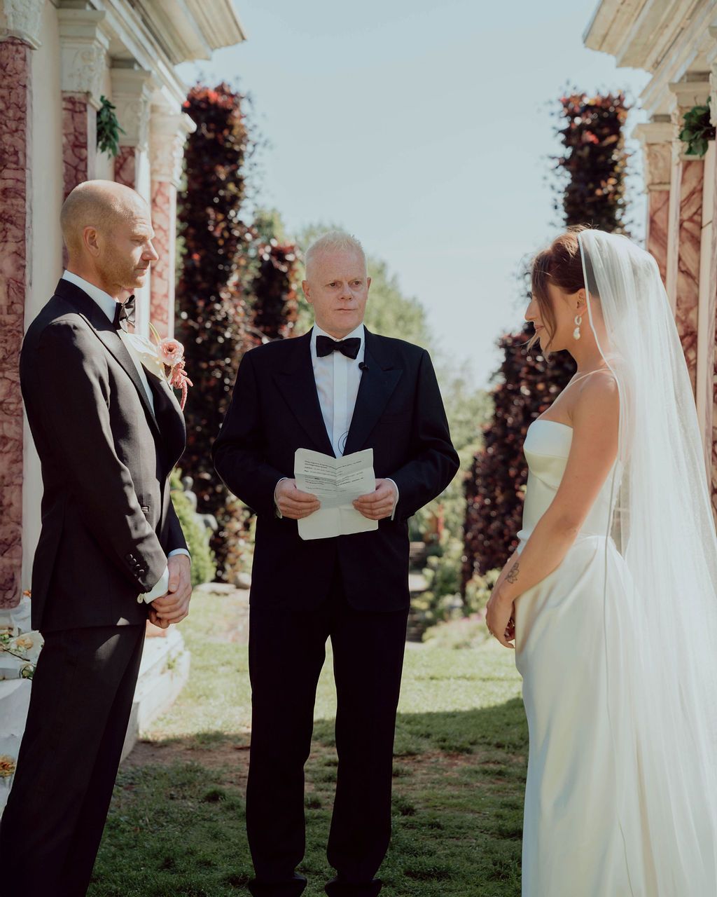 A couple exchanges wedding vows during an outdoor ceremony surrounded by decorative columns and greenery.