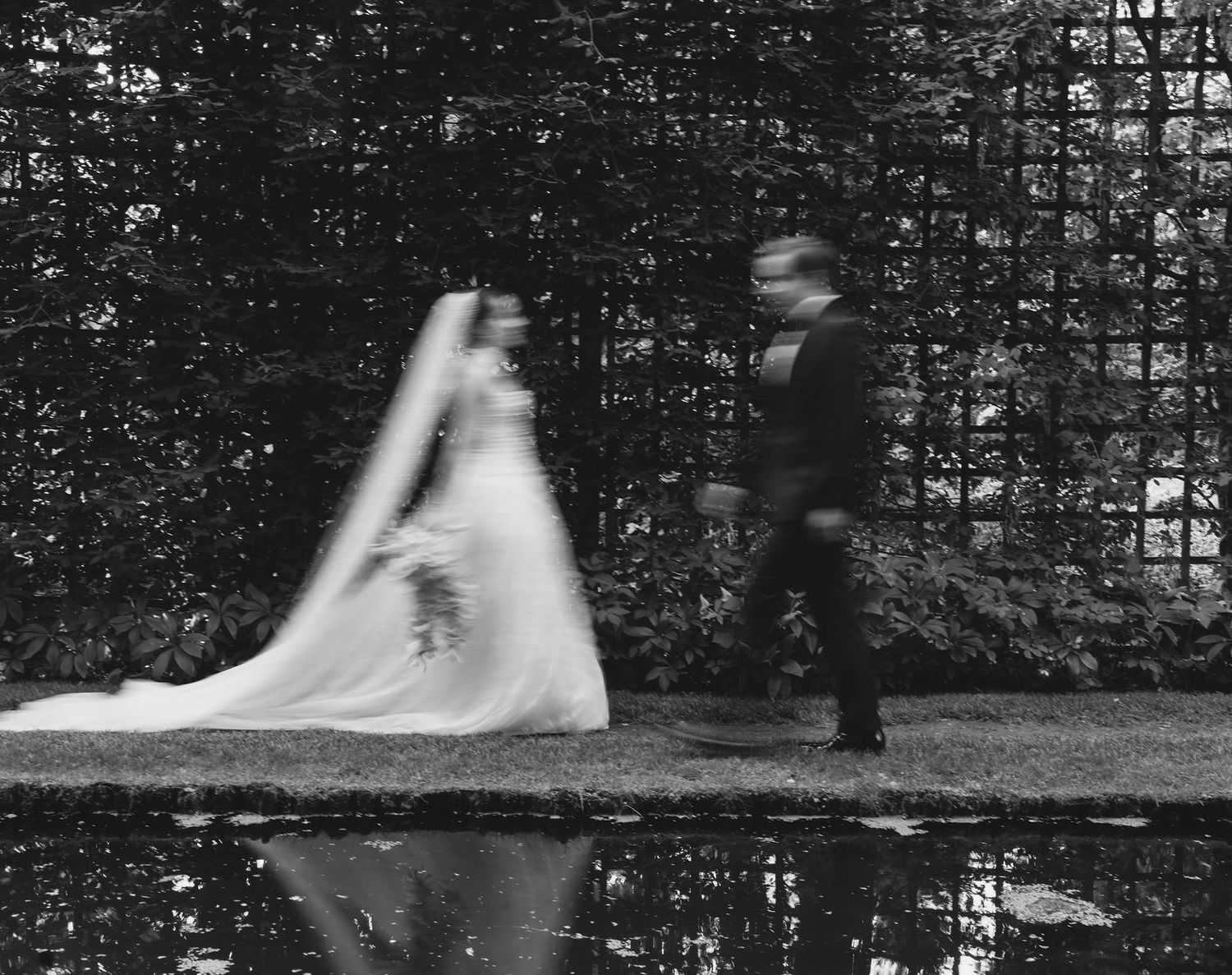 Black and white photo of a bride and groom walking past a reflective pond in a lush garden on their wedding day.
