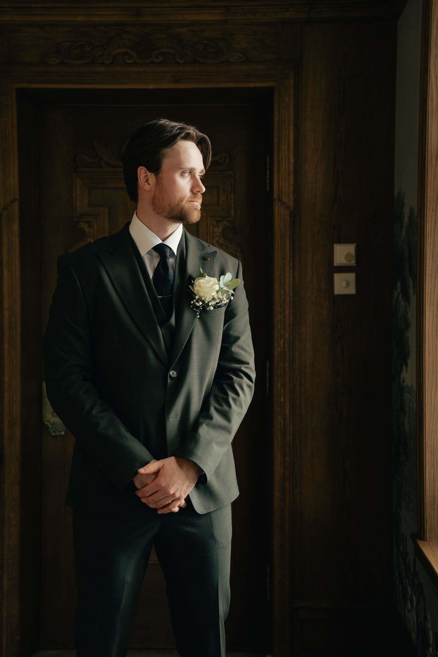 Handsome groom in dark grey suit with floral boutonniere standing in a wooden doorway.