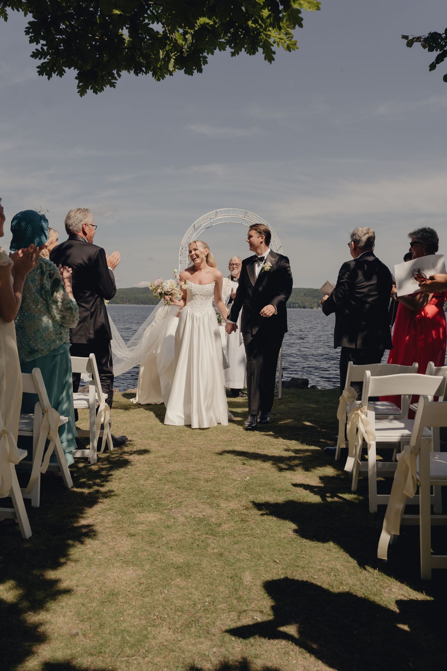 Smiling bride and groom walk down the aisle by the lake as applauding guests stand beside white chairs.