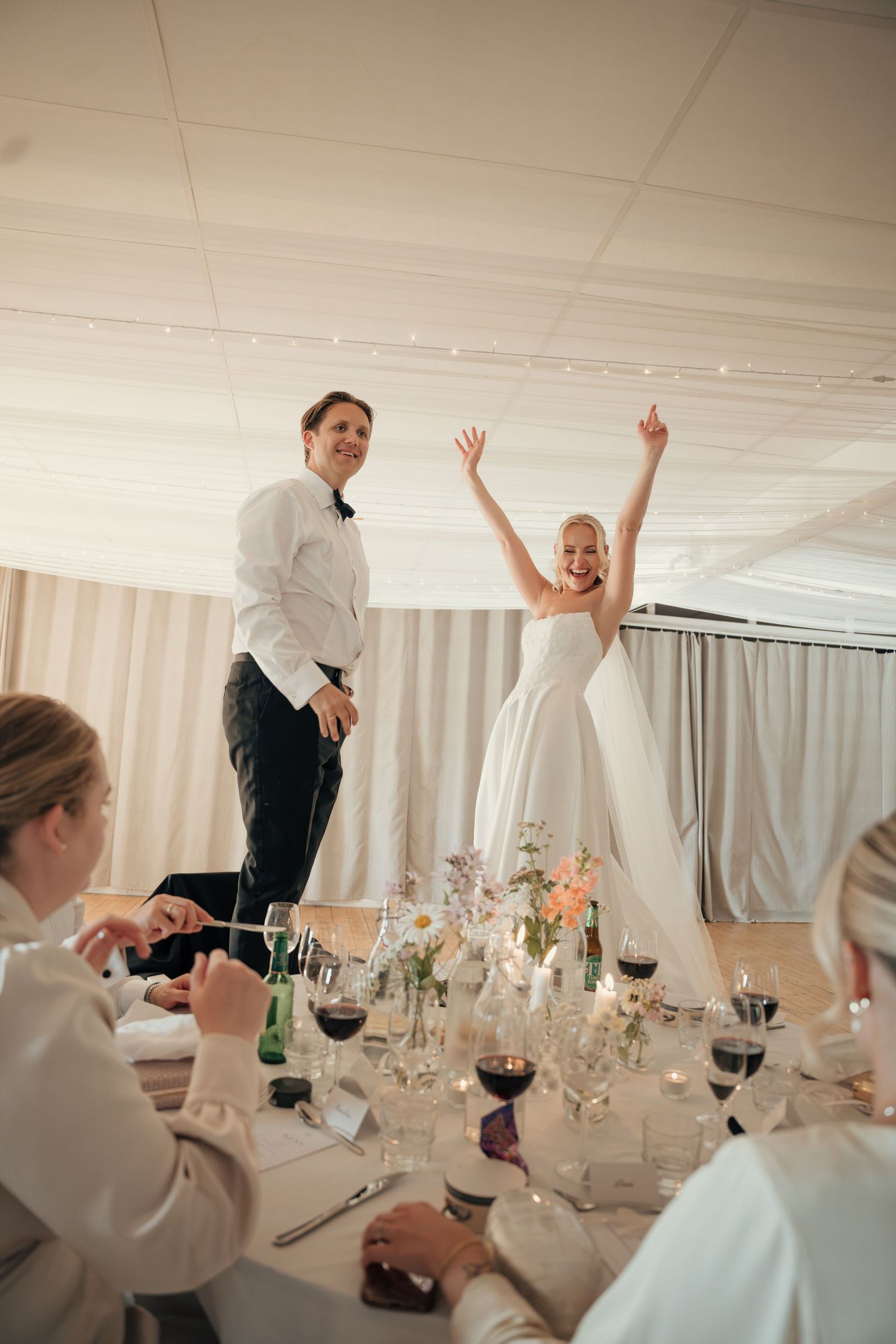Bride with arms raised and groom stand on a reception table celebrating among wine glasses and floral centerpieces.