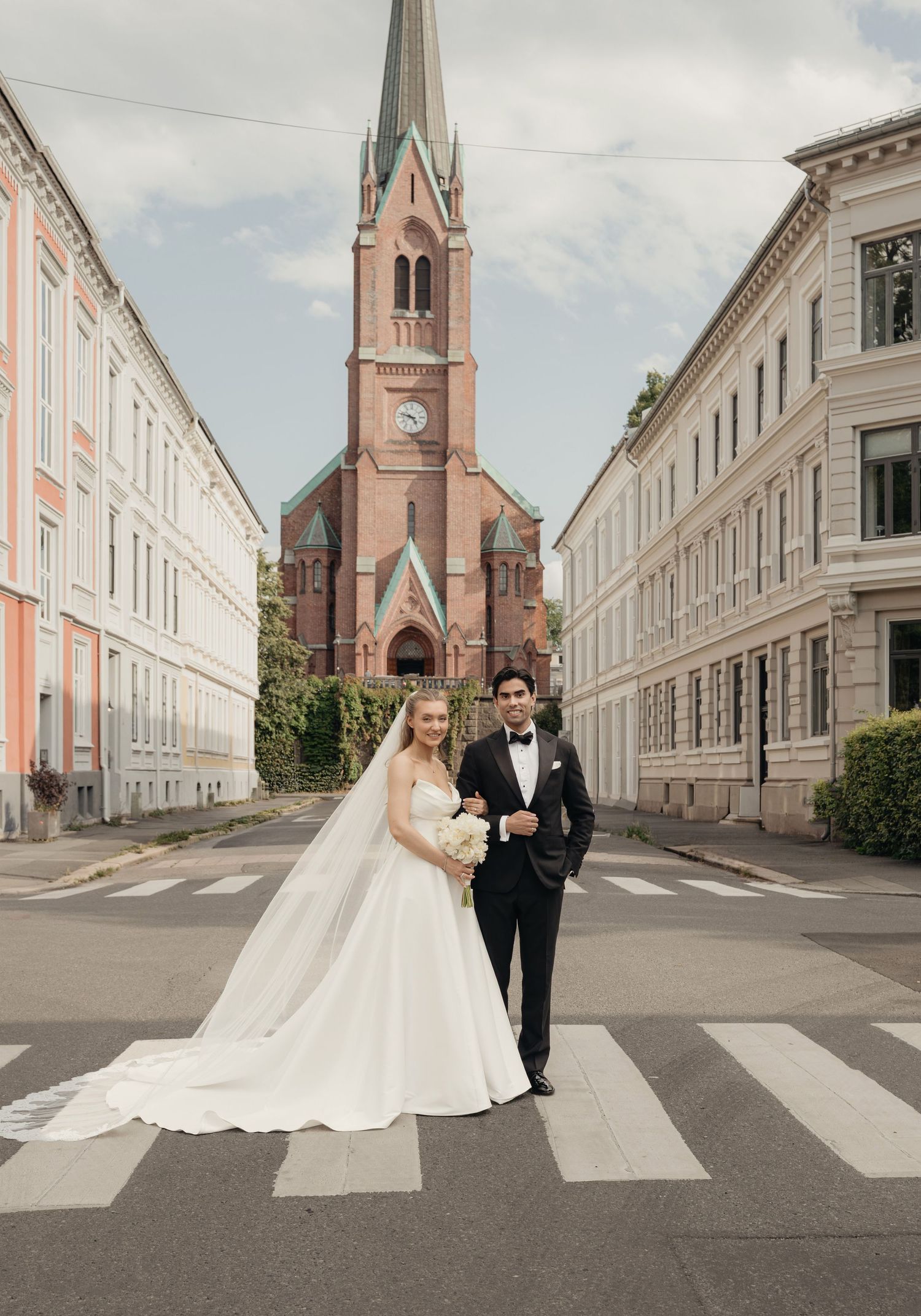 Bride in a flowing white gown and groom in a black tuxedo pose on a crosswalk in front of a red brick church with a tall spire.