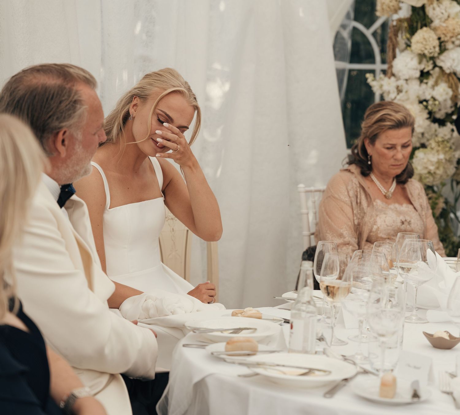 Bride in a white dress wipes tears during an emotional wedding reception speech as guests sit at a elegant table with floral decor.