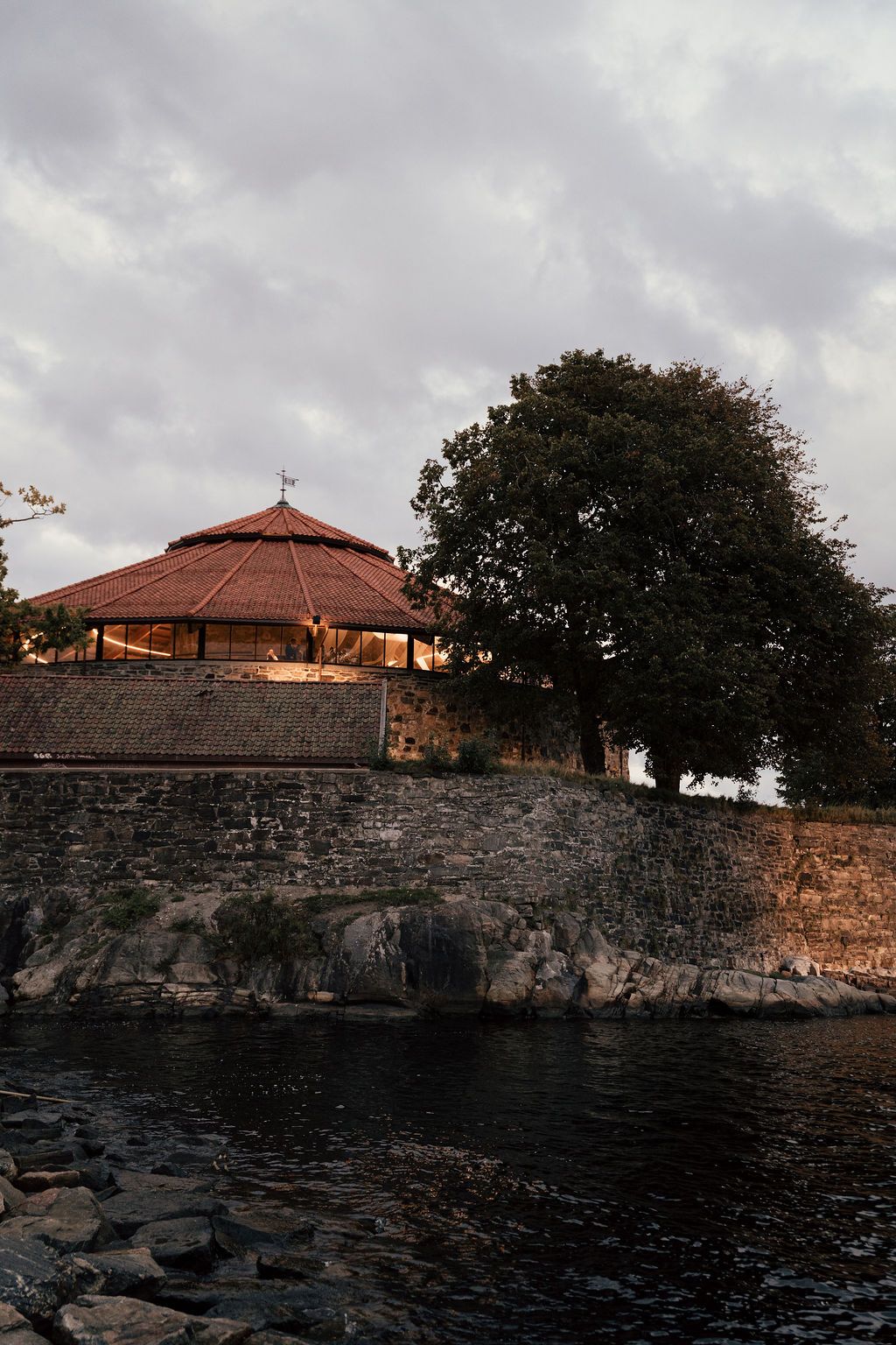 Stone wall and houses with terracotta roofs along a rocky coastline under moody cloudy skies at dusk.