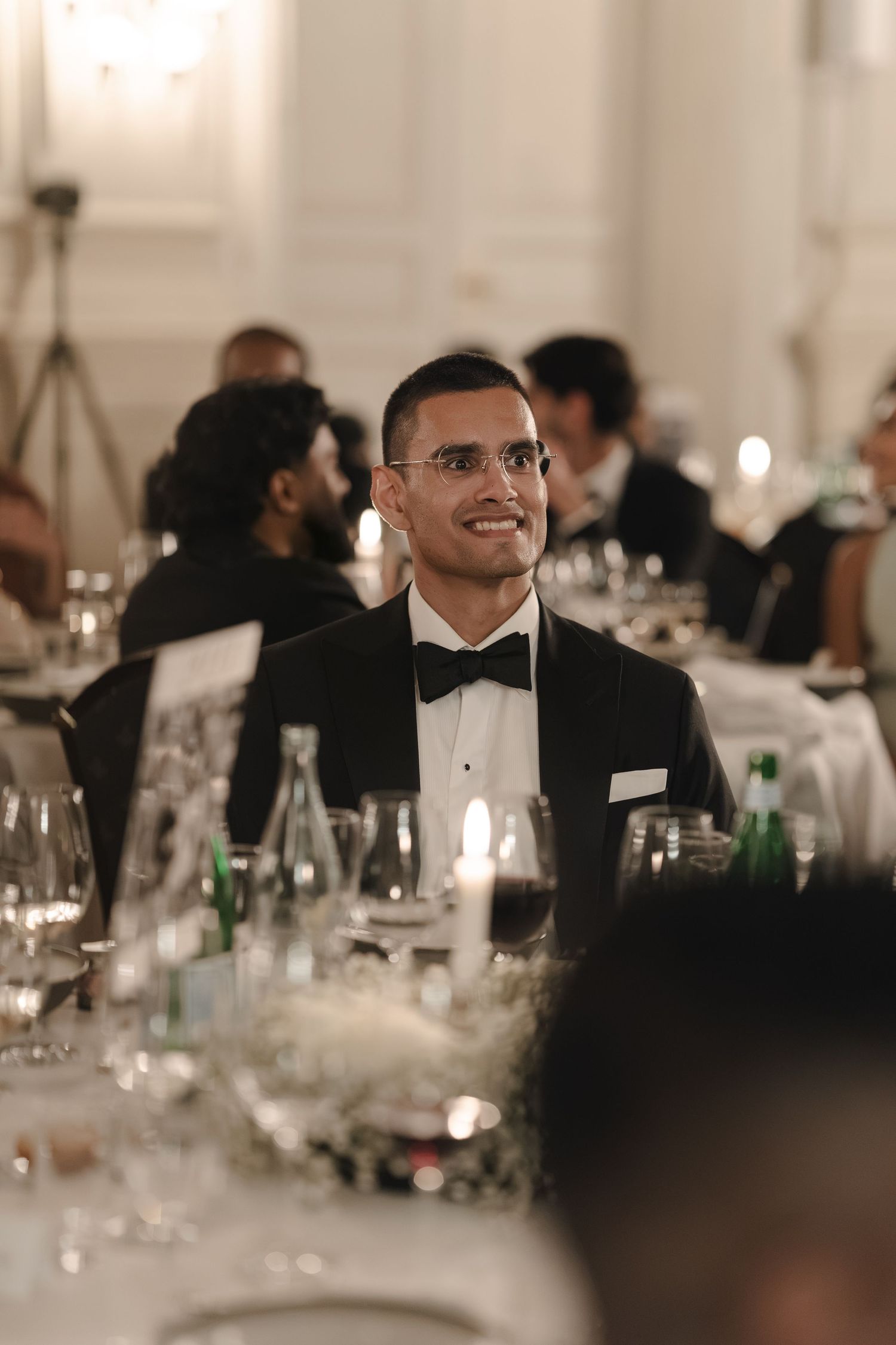A smiling man in glasses, tuxedo and black bow tie sits at an elegant candlelit dinner table with other formal guests behind him.