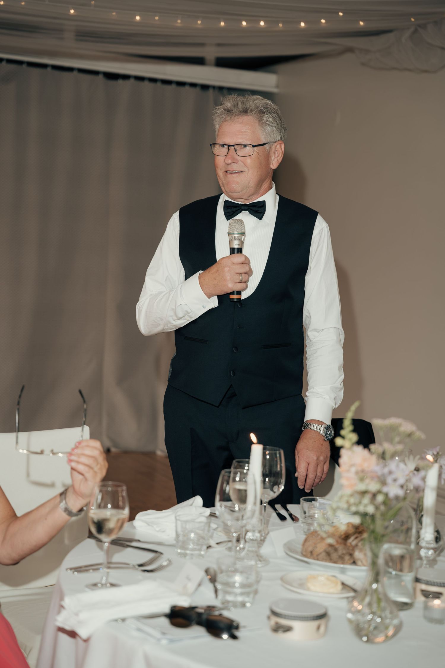 Man in a vest and bow tie holds a microphone while giving a toast at a candlelit wedding reception table.