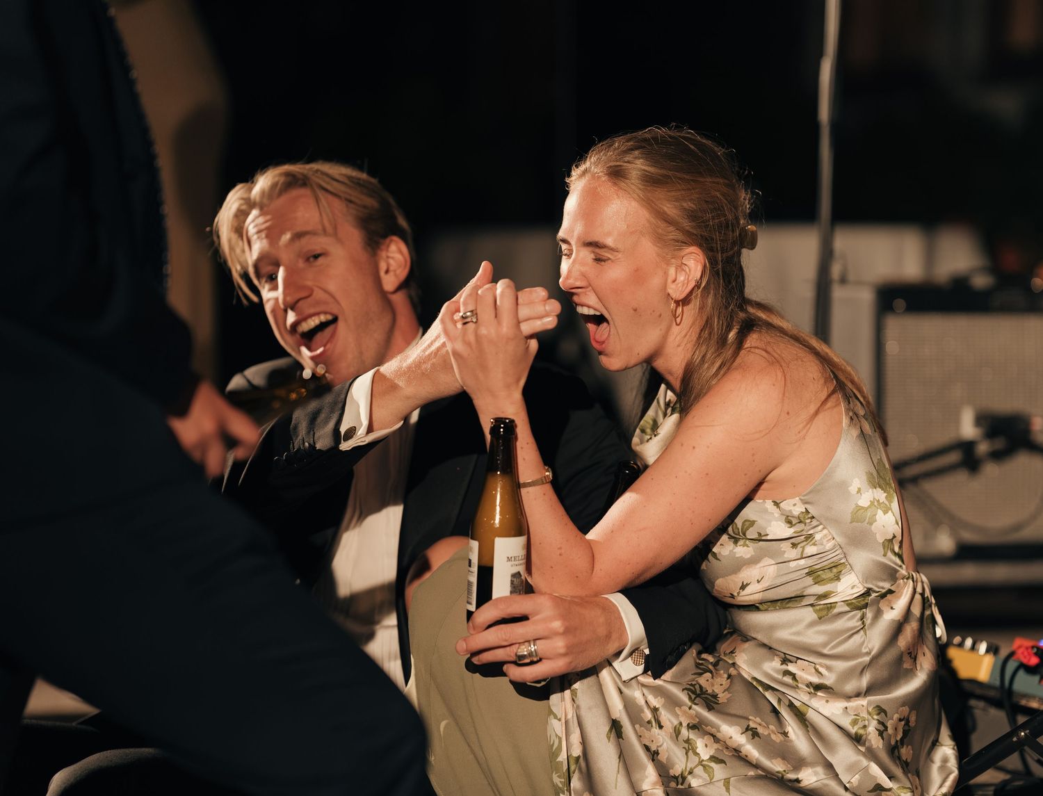 A laughing man and woman in a floral dress share a joyful moment at a party, holding a wine bottle between them.