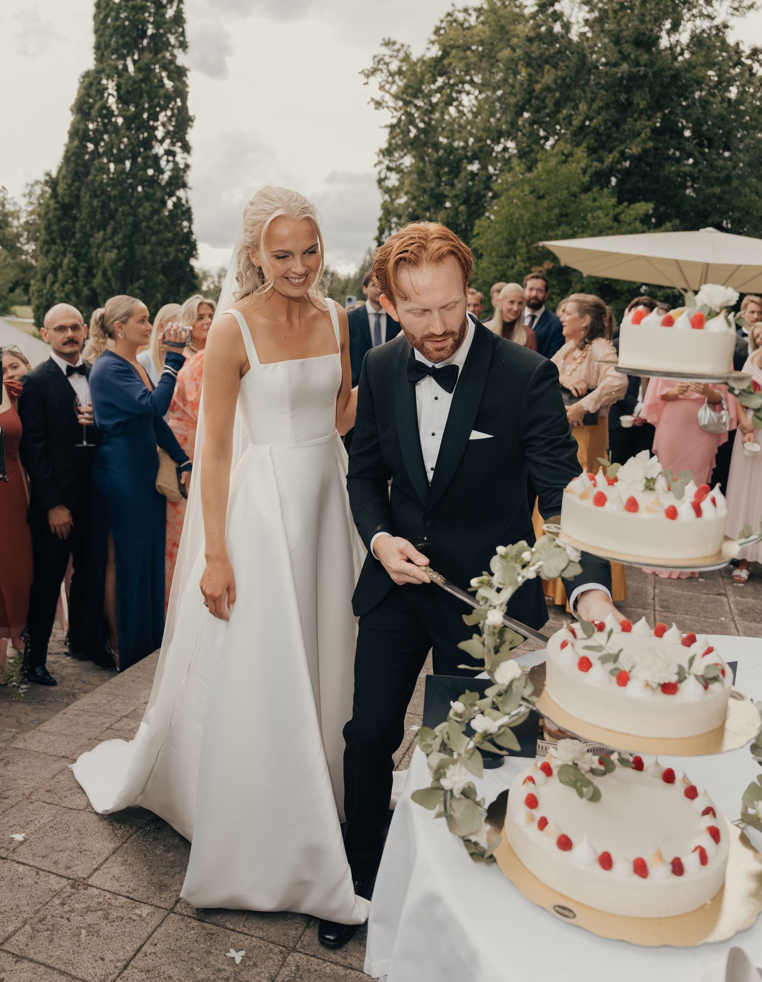 Bride in a white gown and groom in a black tuxedo cut a tiered wedding cake with red berries while guests gather outdoors.
