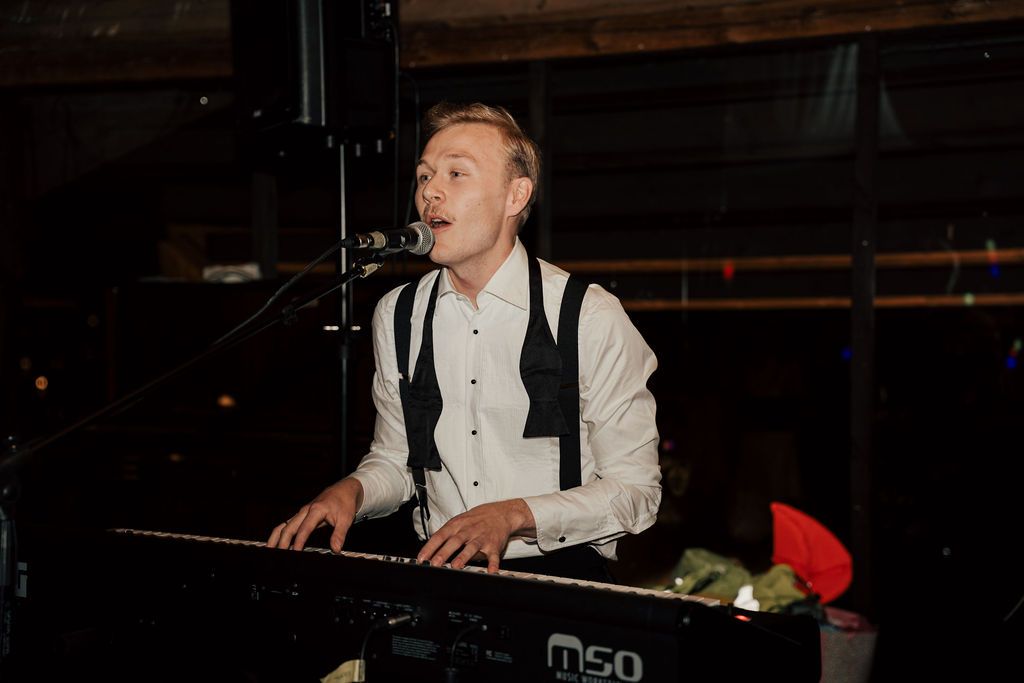 A musician performs enthusiastically behind electronic music equipment on a dark stage.