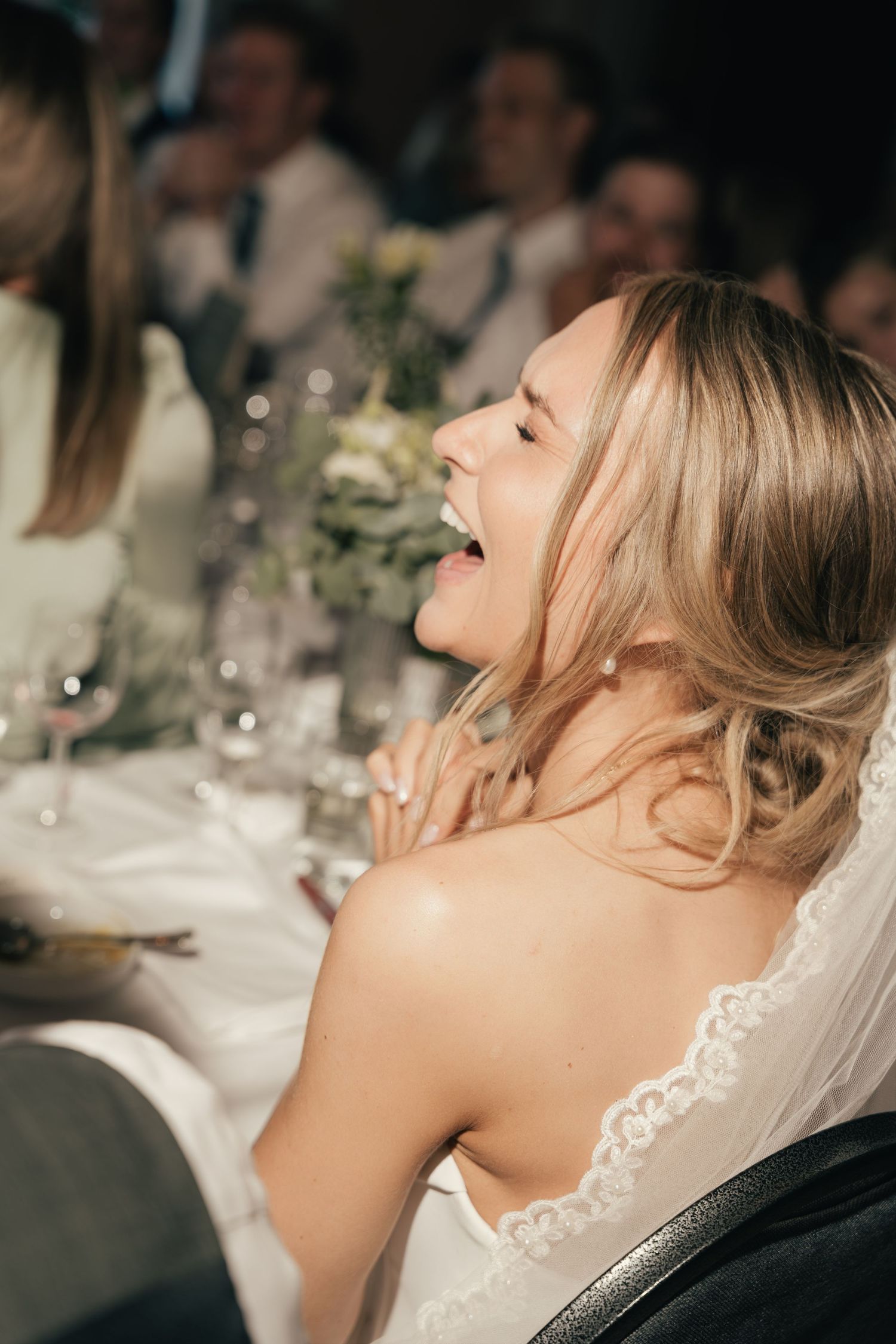 Joyful blonde bride laughing at wedding reception table wearing strapless lace wedding gown.