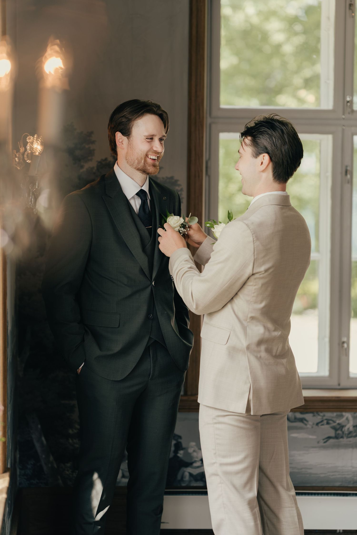Groomsman in beige suit pinning boutonniere on groom in black suit near window at wedding.