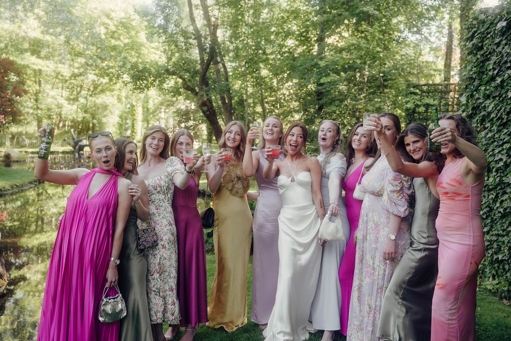 A group of people in pink and white formal attire stand together for a wedding photo in an outdoor garden setting.