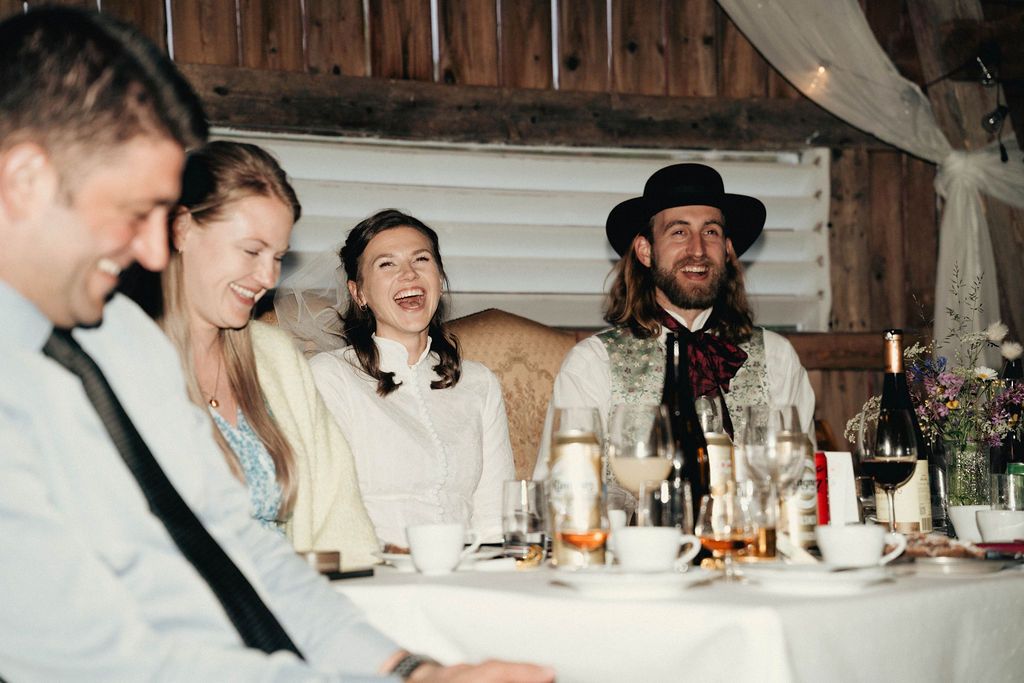 Wedding guests seated at long tables in a rustic barn setting with candles and floral centerpieces during dinner service.
