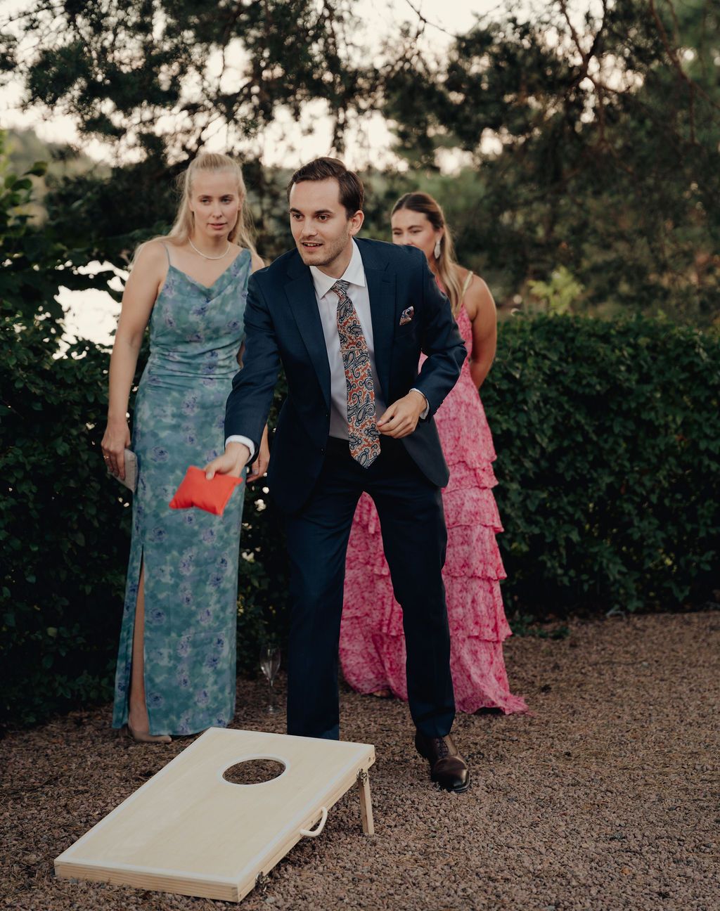 Wedding guests playing cornhole outdoors at a garden reception with people in formal attire.